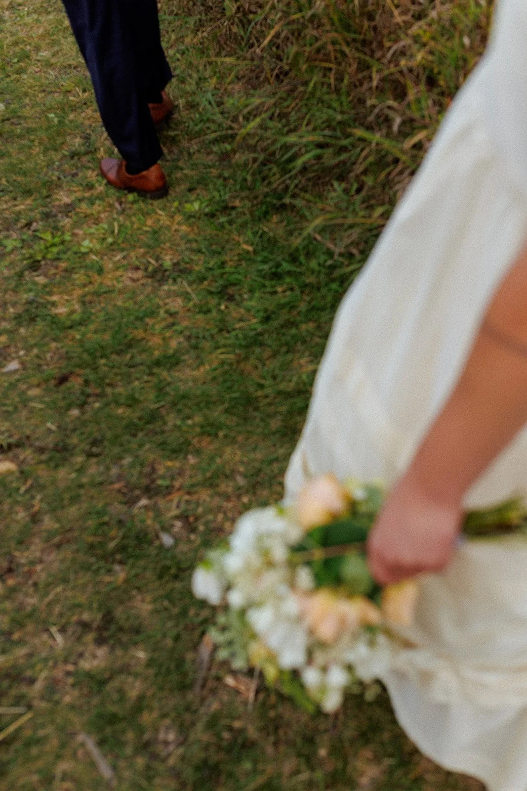 A person dressed in white holding a bouquet of flowers, standing on grass, with a man in dress shoes and navy pants standing nearby on the grass.