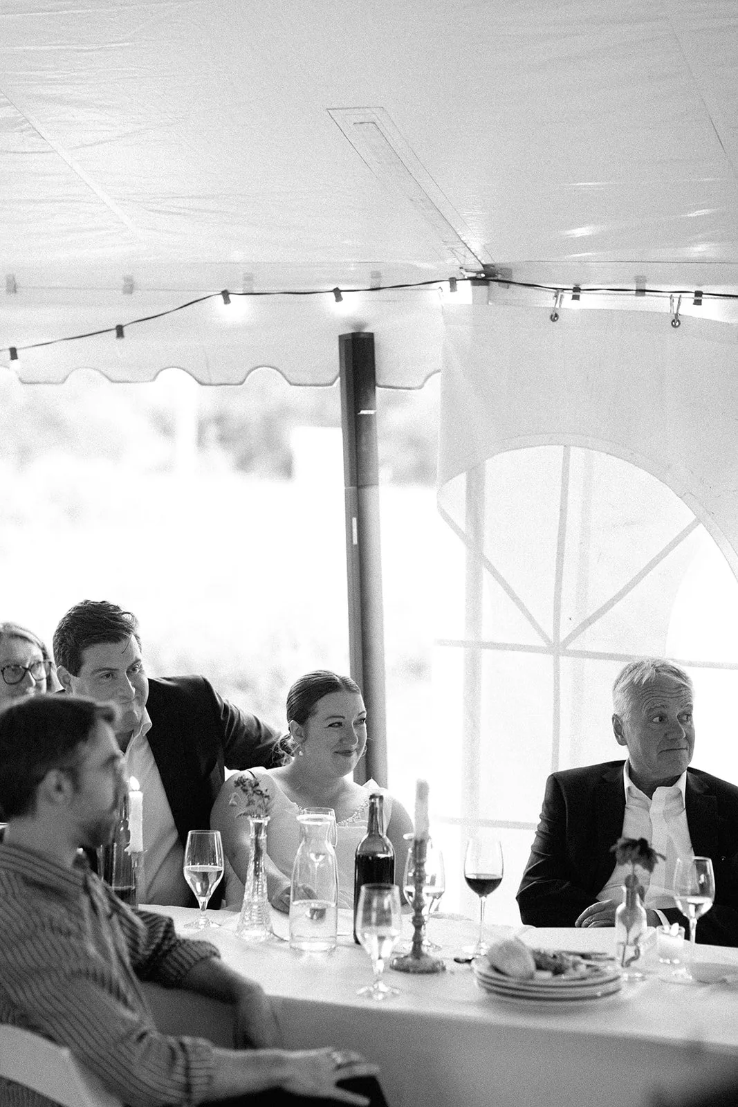 People sitting at a table during a formal event or celebration, with drinks and a plate of bread in front of them, inside a tent or pavilion with string lights and large windows.