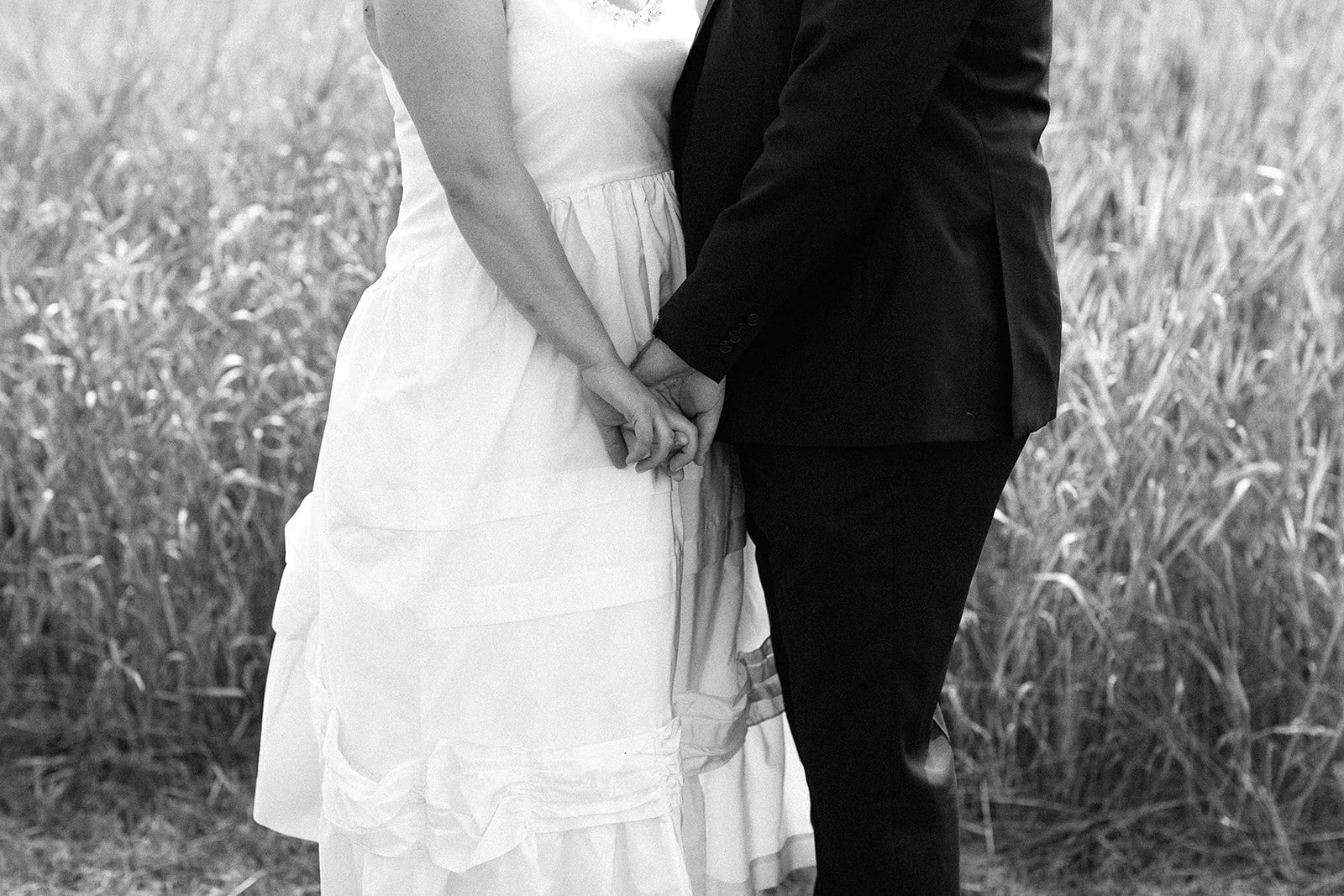 A black and white photo of a bride and groom holding hands, standing in a field of tall grass.