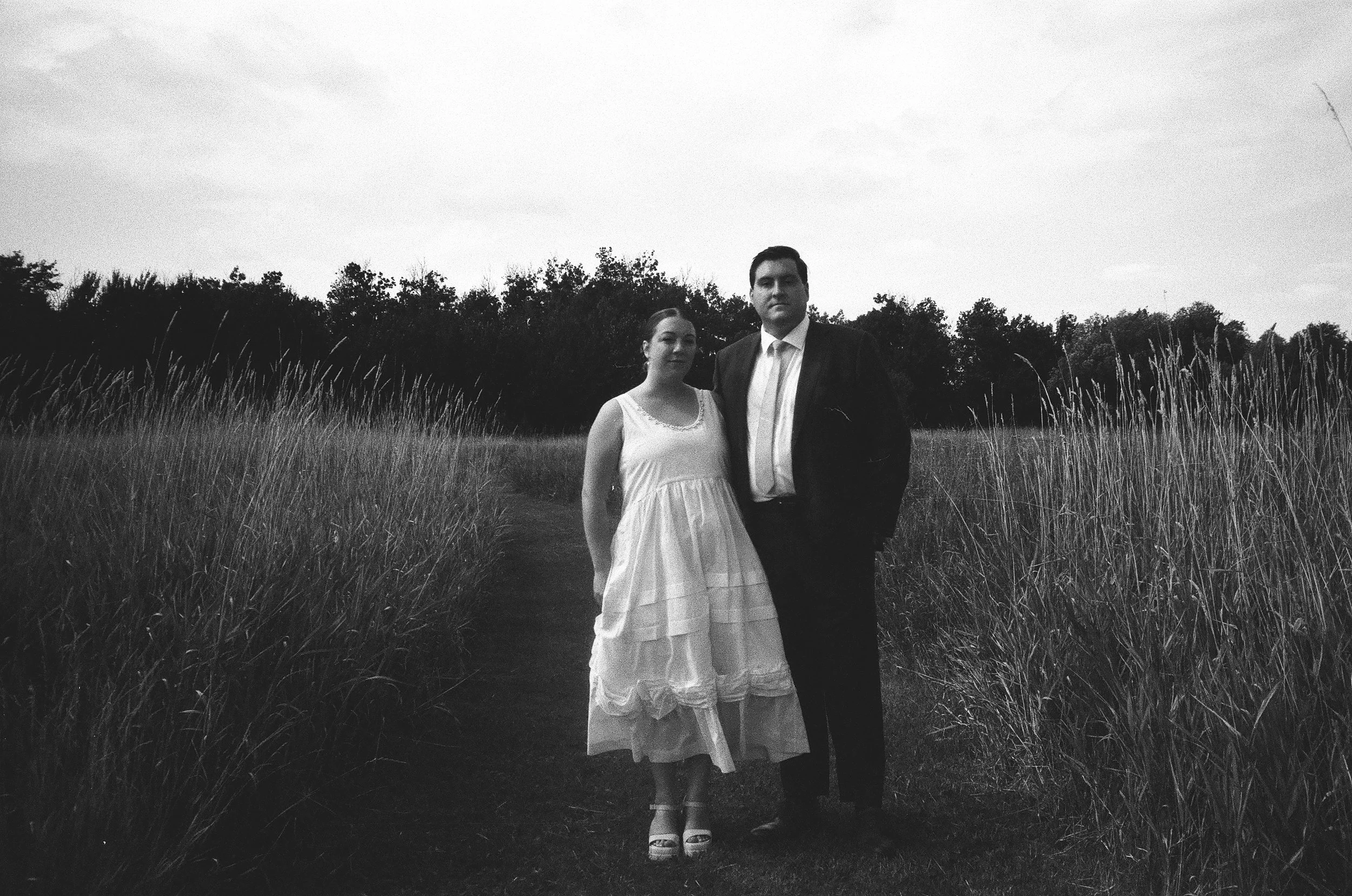 A black and white photo of a man and woman standing together on a narrow dirt path in a grassy field, with trees in the background.