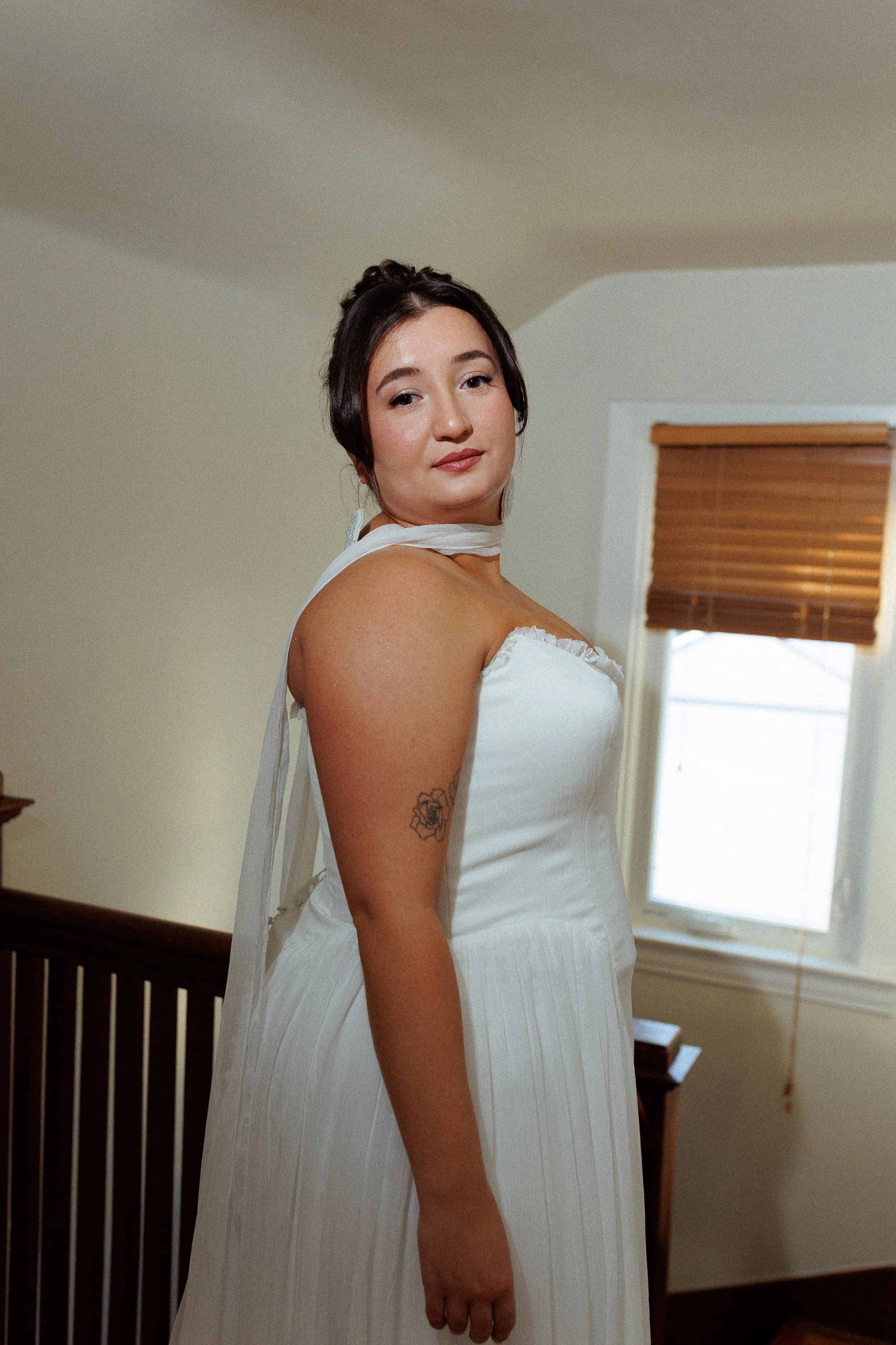 A woman with dark hair styled in an updo wearing a white dress, standing indoors near a window with brown blinds, looking at the camera.