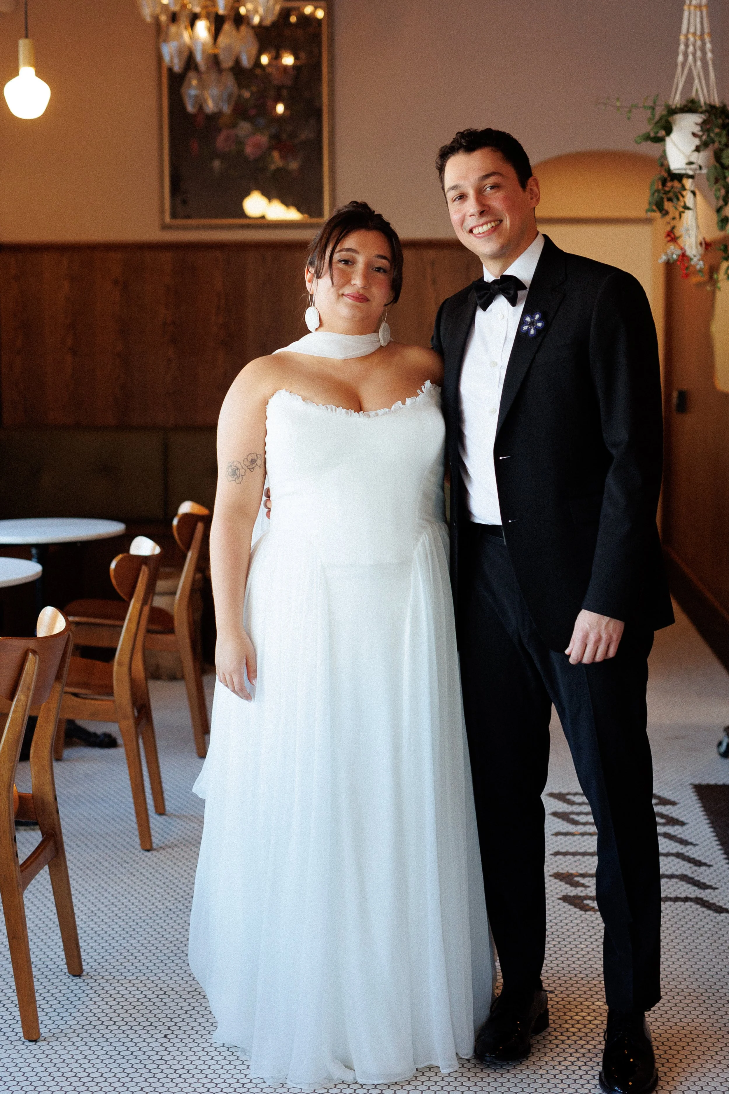 A bride and groom standing together indoors, smiling. The bride is wearing a white wedding gown with a choker, and has tattoos on her arm. The groom is dressed in a black tuxedo with a bow tie.