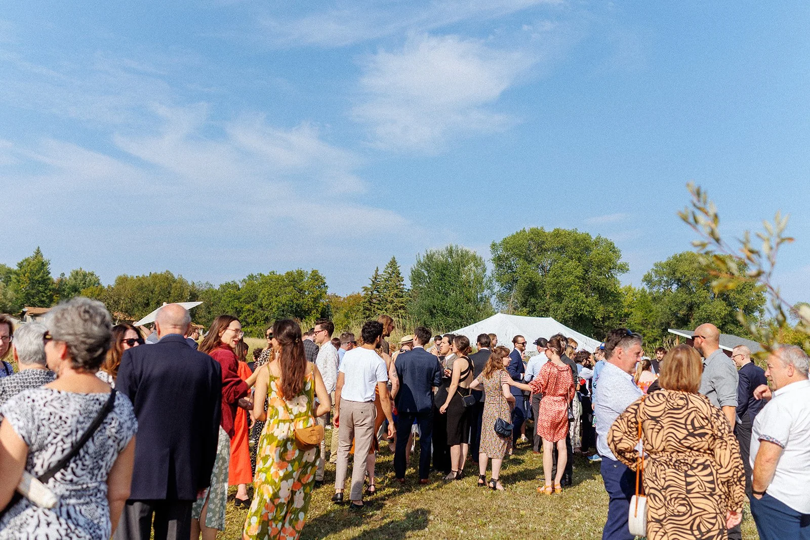 A large outdoor gathering of people at a social event on a sunny day, with trees and a tent in the background.