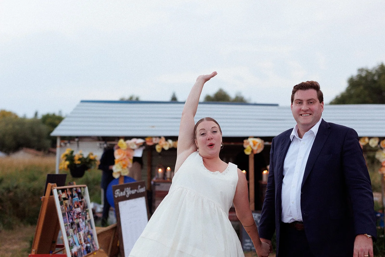 Couple at outdoor wedding reception, woman in white dress with raised arm, man in suit next to her, decorated wooden structure with flowers in background.