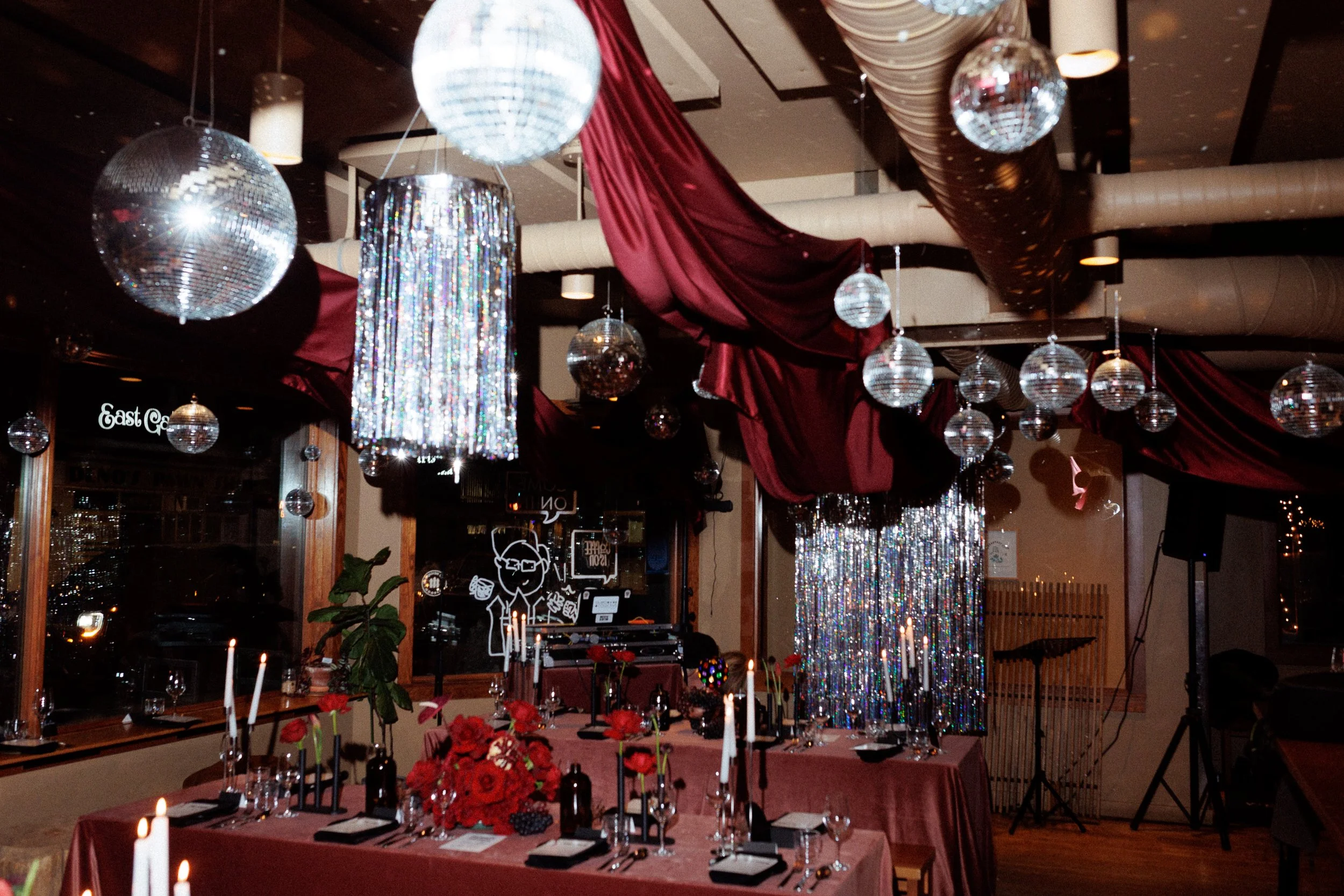 Decorated interior party venue with hanging disco balls, large red drapes, crystal bead curtains, a table with red flowers, candles, wine glasses, and place settings.