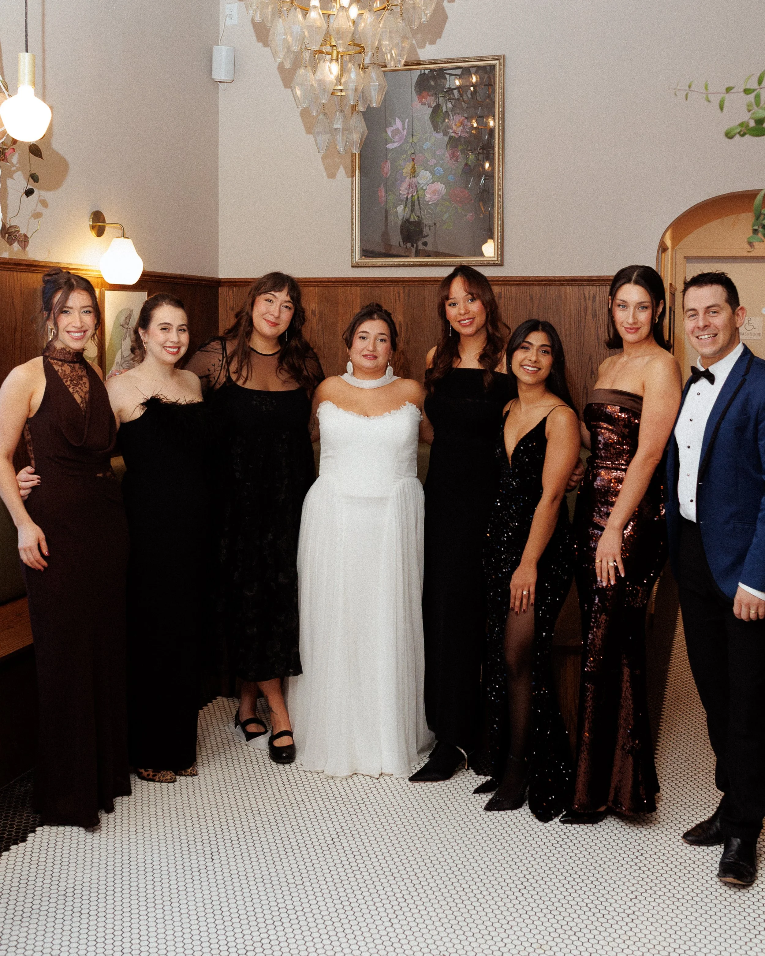Group of nine people dressed in formal attire, standing in a row inside a decorated room with wood-paneled walls, framed artwork, and a chandelier.