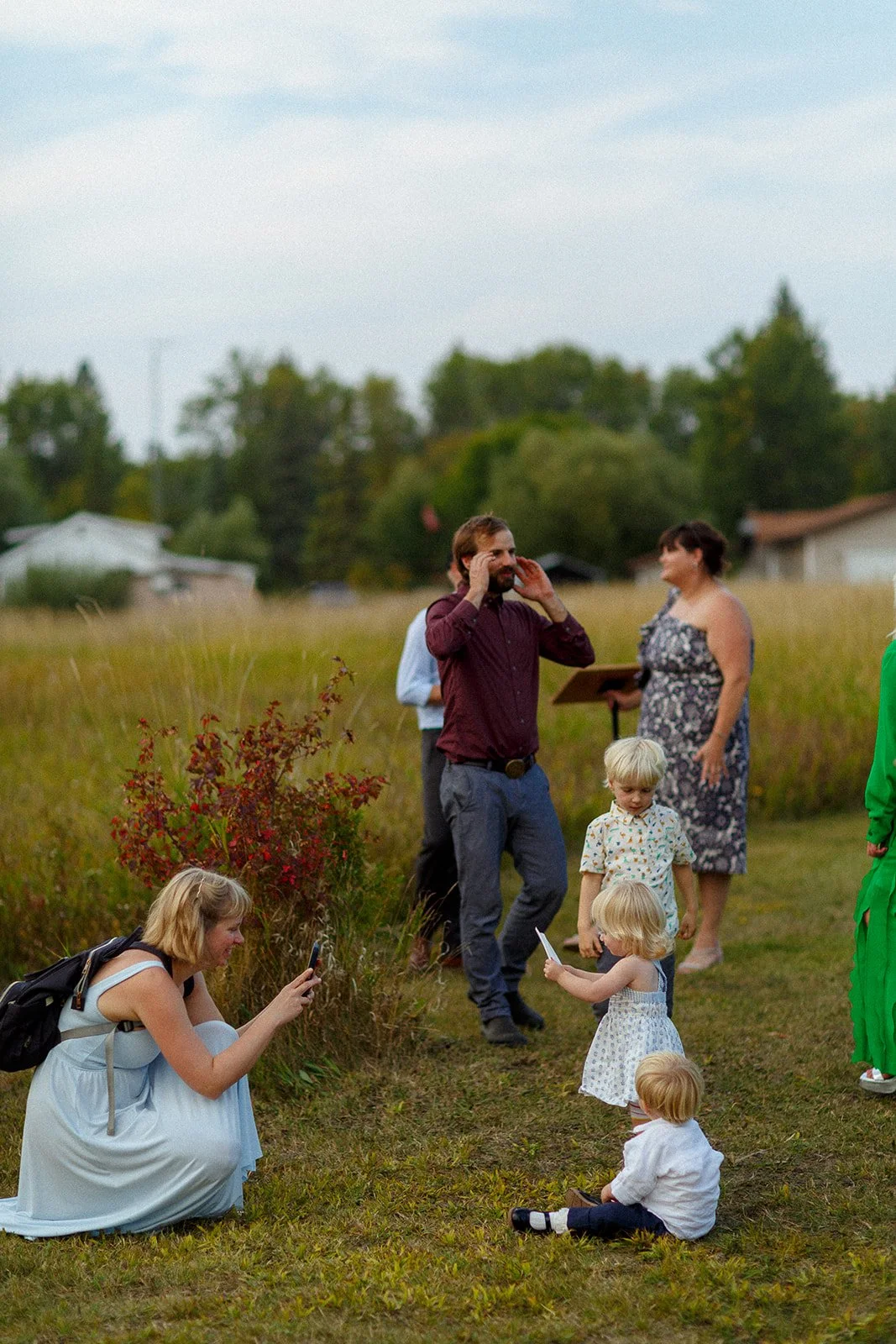 People gathered outdoors in a grassy field, some taking photos and others engaging with children, with a few houses and trees in the background.