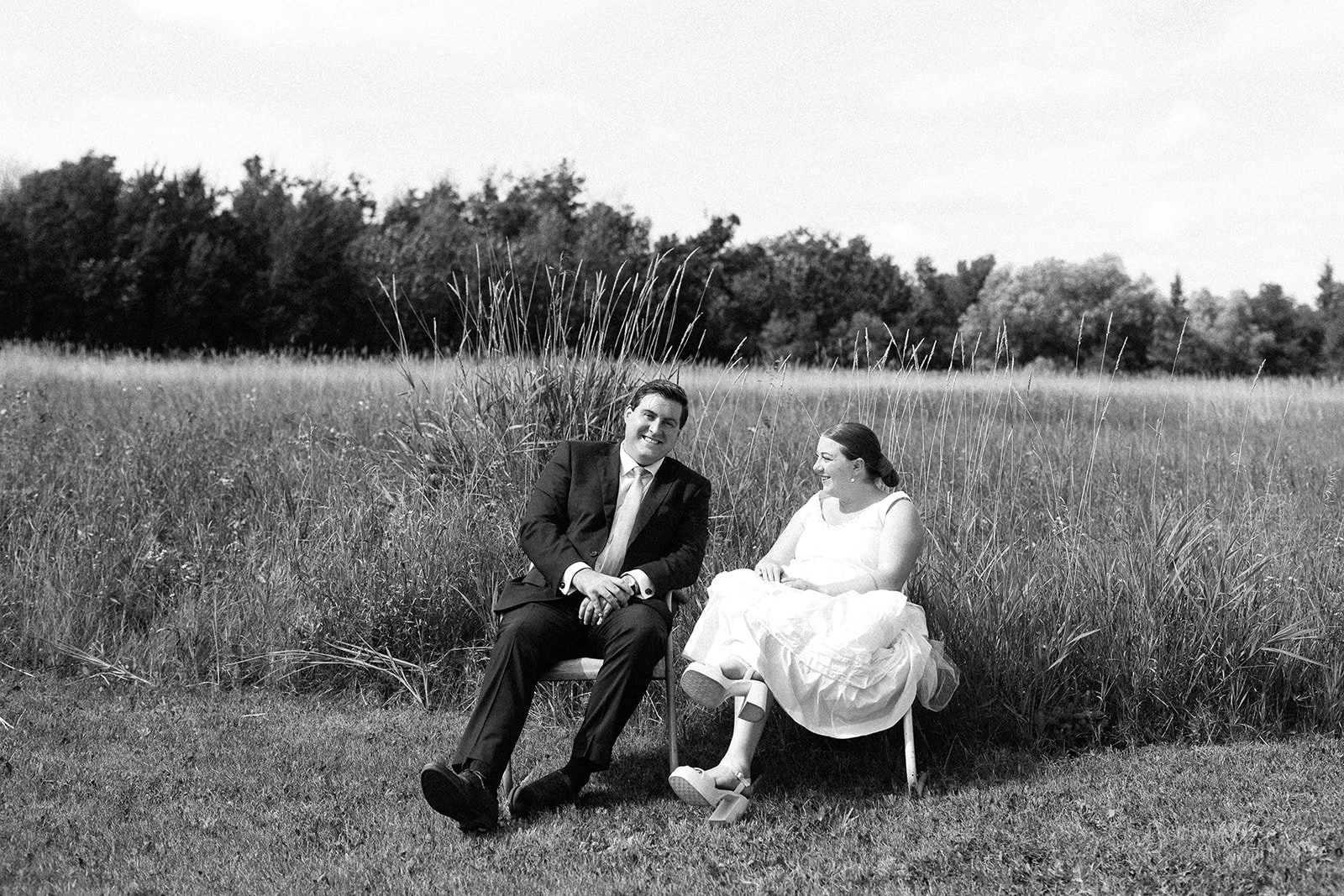 Black and white photo of a man and woman sitting on chairs outdoors in a grassy field, smiling and looking at each other, with tall grass and trees in the background.