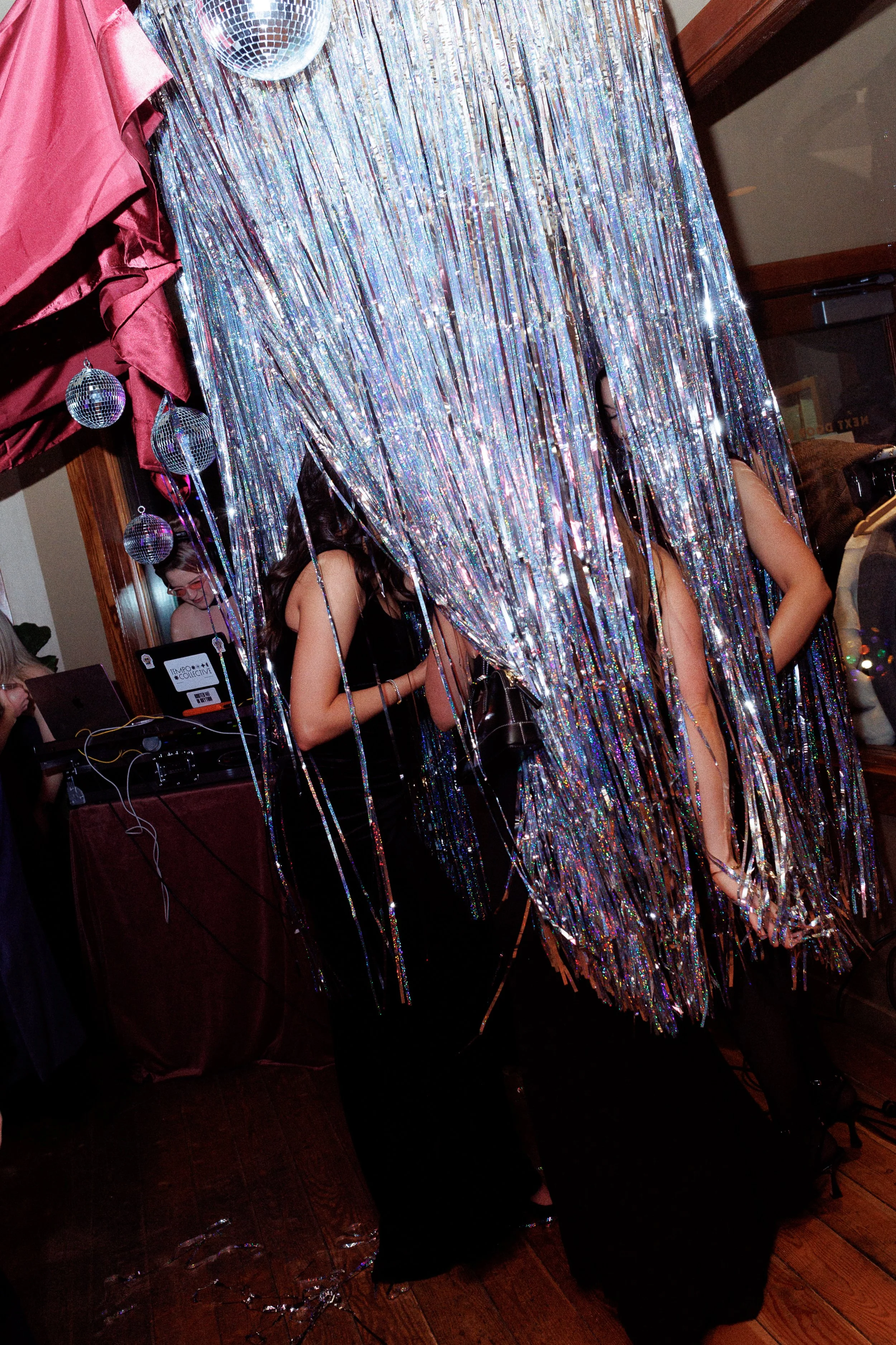 People dancing behind a shimmering silver tinsel curtain at a party with disco balls and a DJ booth.