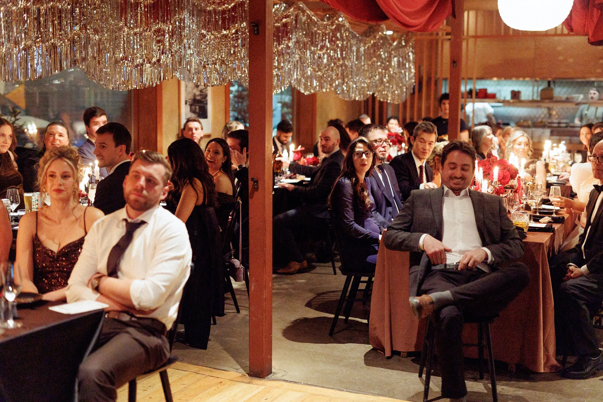 Guests at a formal dinner event seated at tables decorated with candles and red floral centerpieces, under hanging golden tinsel decorations.