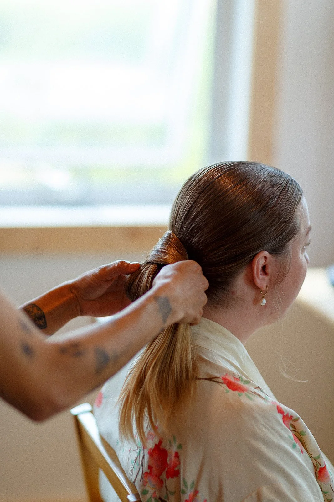 A woman with brown hair is getting her hair styled by a person with tattoos on their arms, in a well-lit room with a window in the background.