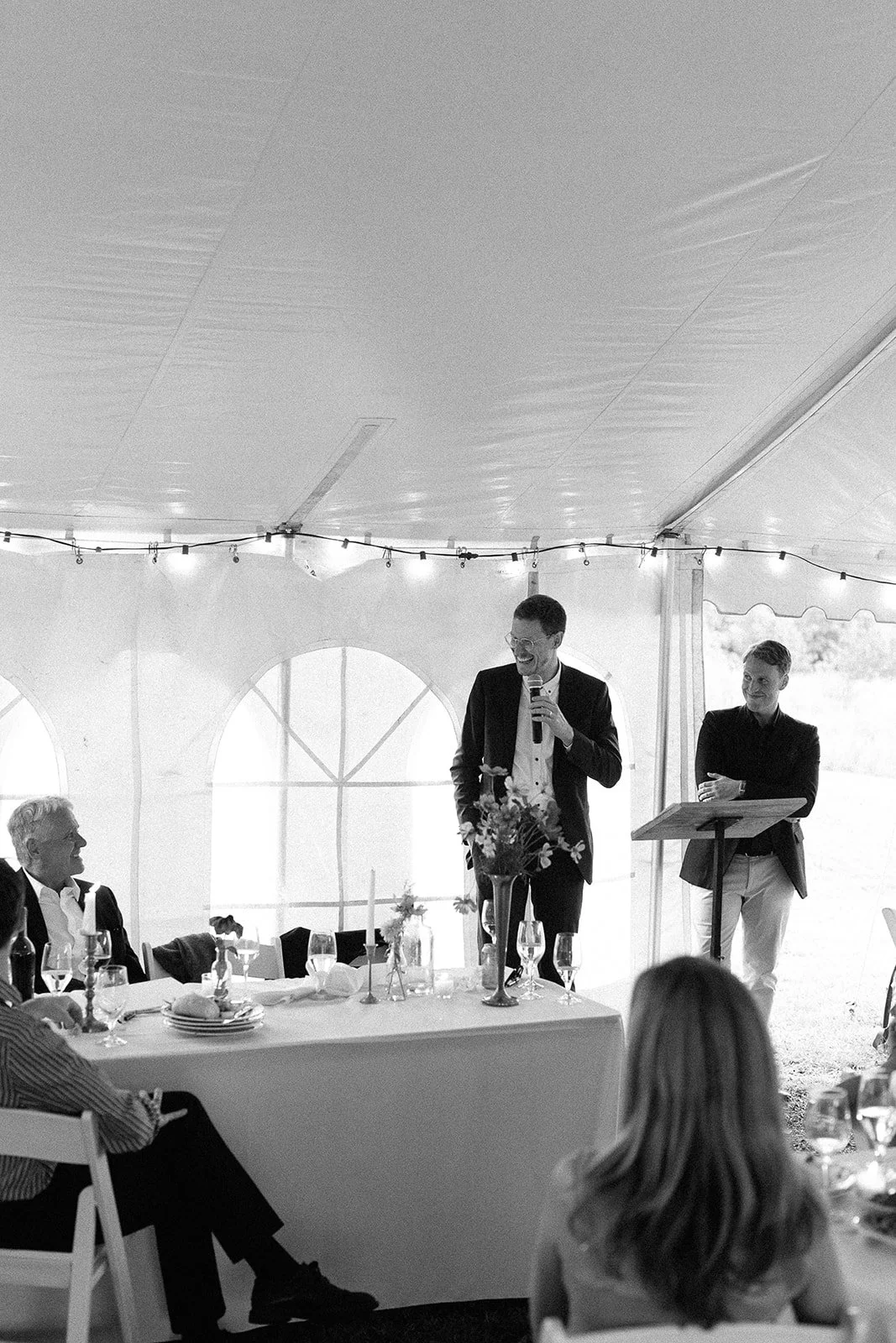 Black and white photo of a wedding reception under a large tent, with a man giving a speech at a podium decorated with flowers, surrounded by seated guests.
