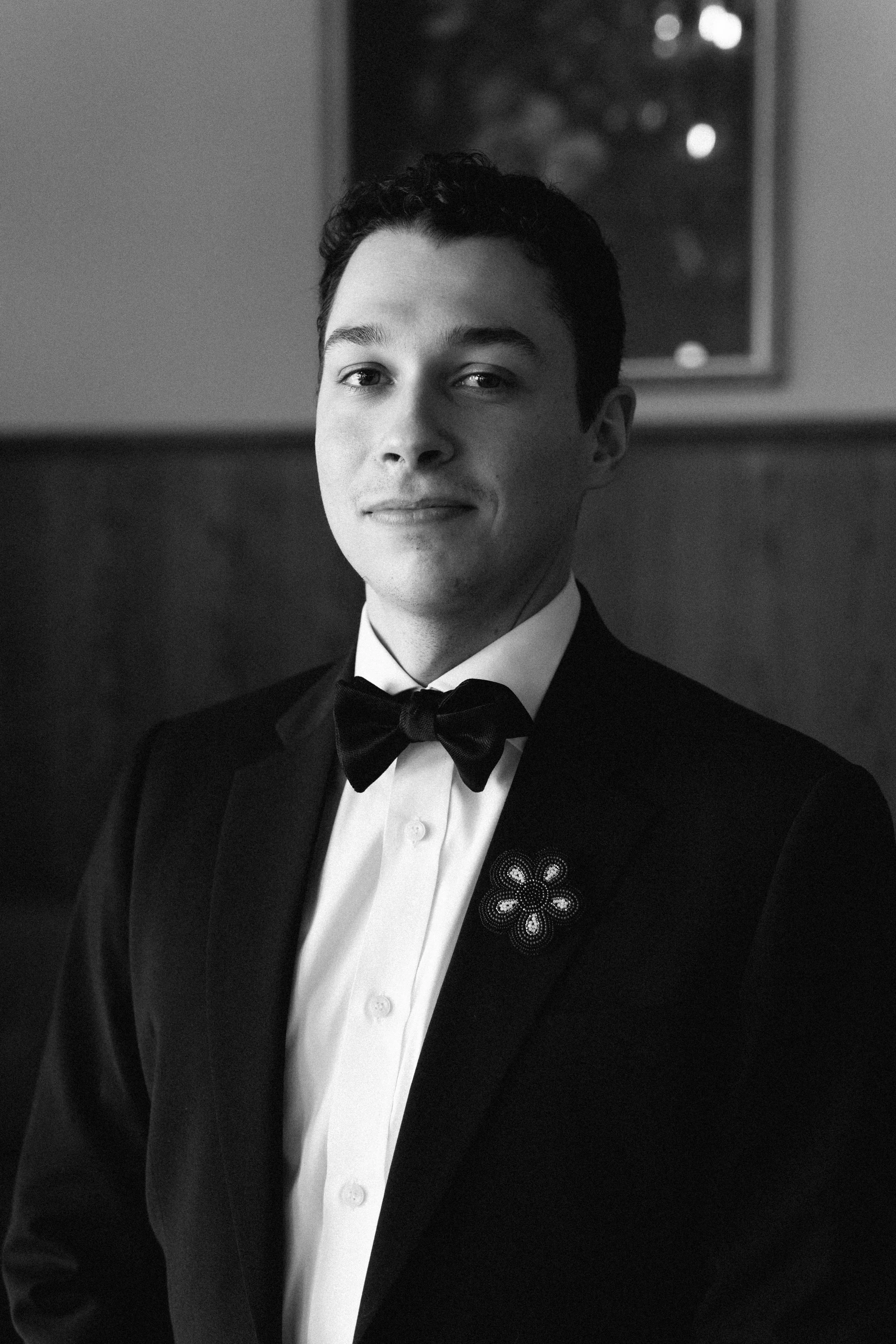 Black and white portrait of a young man in a tuxedo with a bow tie, standing indoors with a window in the background.