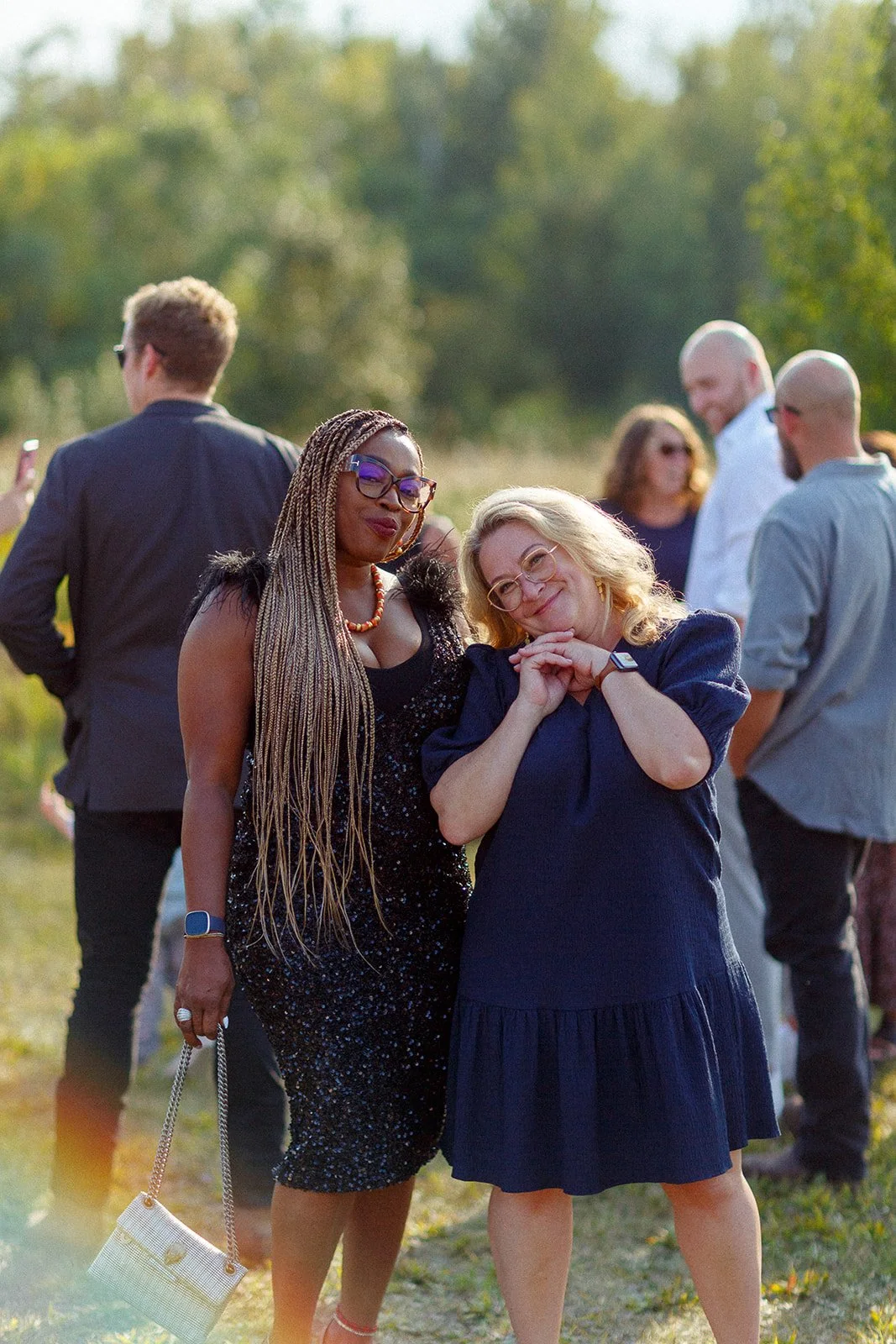 Two women standing close together outdoors in a grassy area during a sunny day. The woman on the left has long braided hair, glasses, a black sequined dress, and is holding a white purse. The woman on the right has blonde hair, glasses, a blue dress,