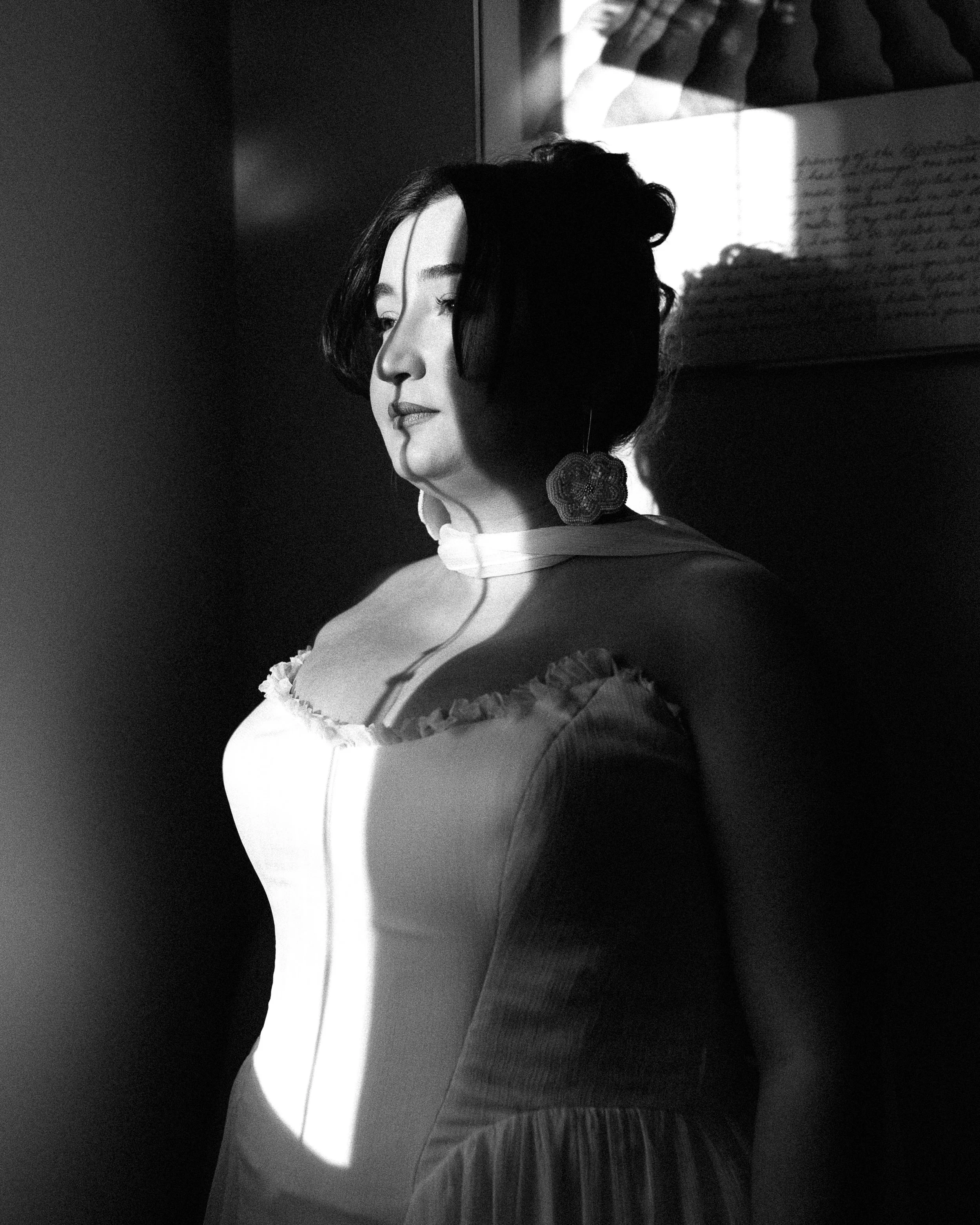 Black and white portrait of a woman with short dark hair, wearing a dress with ruffled details and large earrings, standing against a wall with a window.