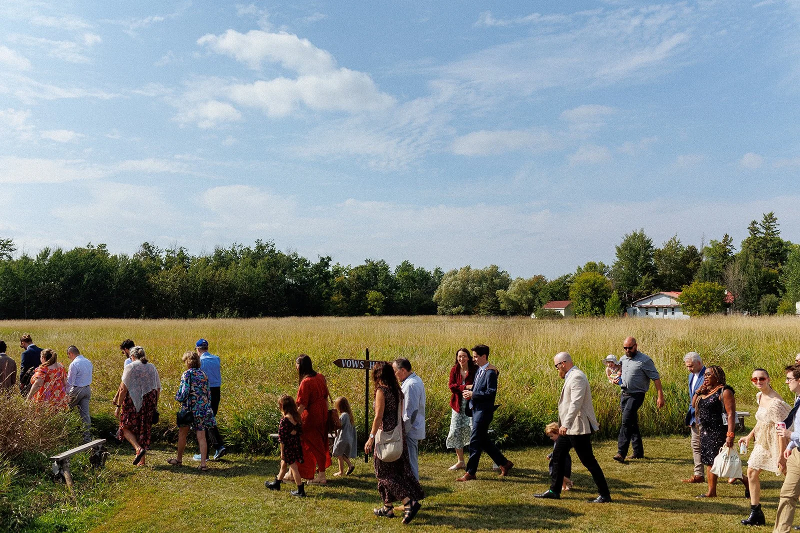 A group of people dressed formally walking through a grassy field, with a signpost labeled 'VOWS' nearby, on a bright day with a partly cloudy sky and trees in the background.