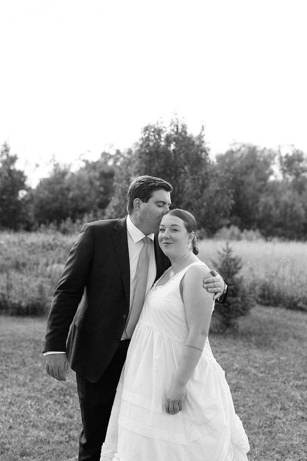 Black-and-white photo of a man in a suit kissing a woman in a white dress on the forehead outdoors, with trees and grass in the background.