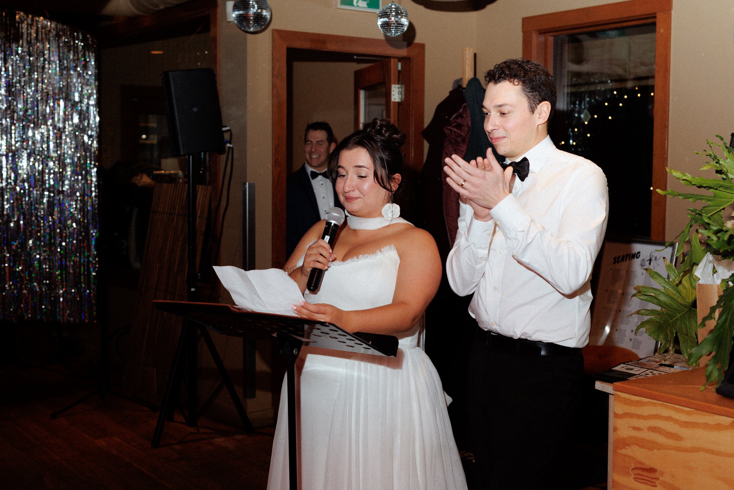 A bride reading a speech while holding a microphone, with a groom clapping beside her, at a wedding reception, with guests and decorative elements in the background.