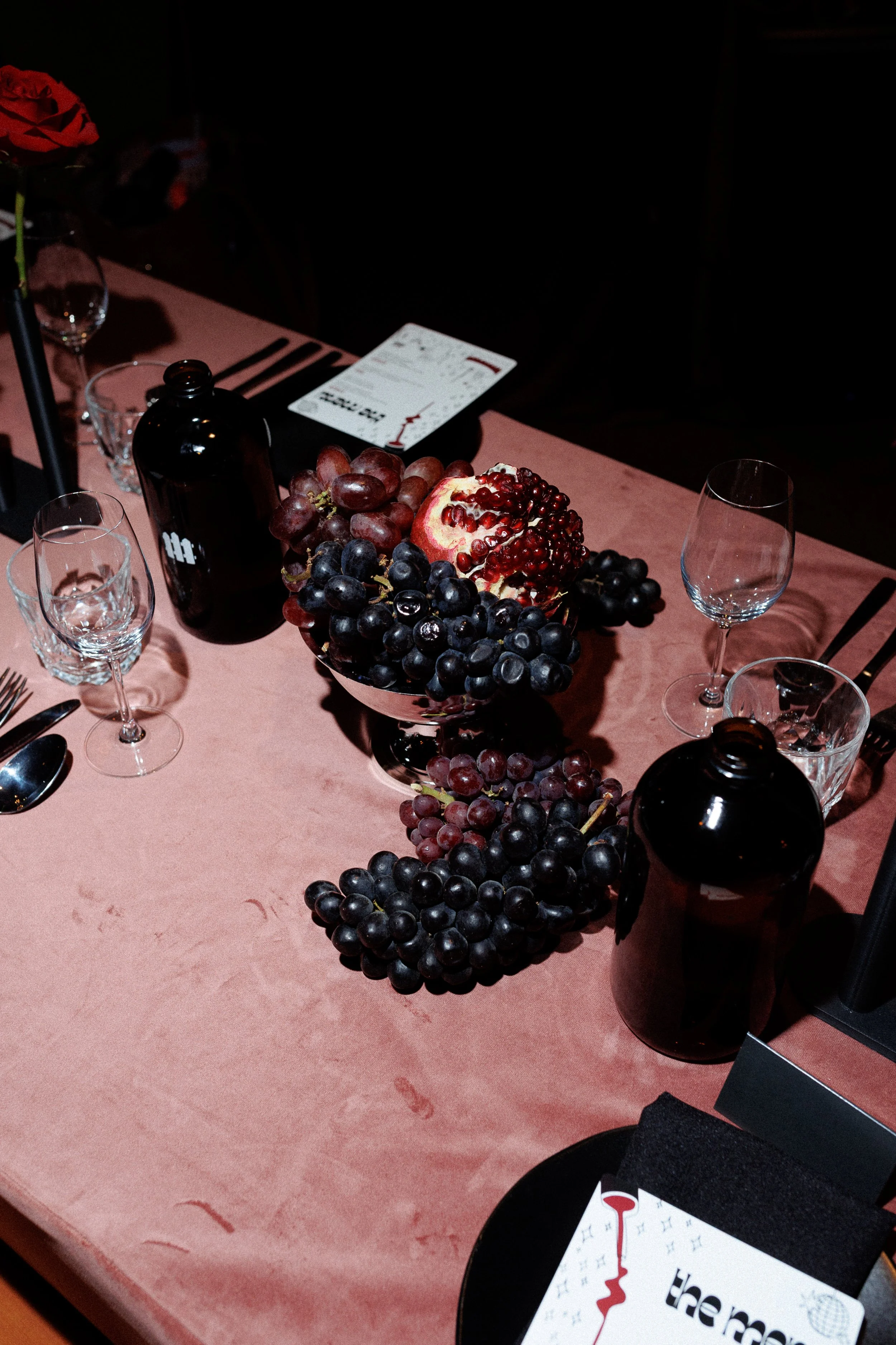 A dining table with a pink tablecloth, decorated with a bowl of grapes and pomegranate, surrounded by wine glasses, black bottles, and tableware.