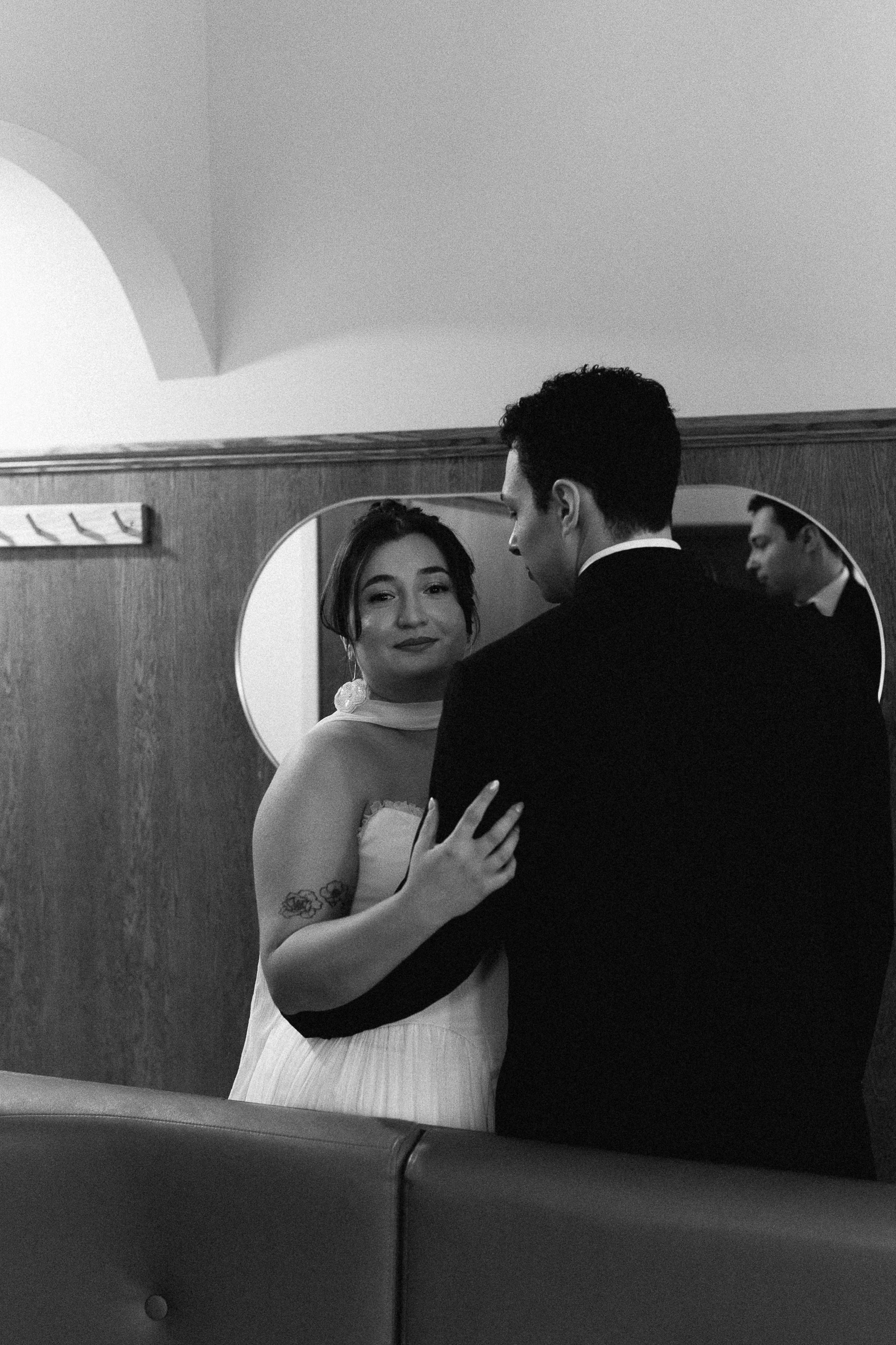 A black and white photo of a couple in formal attire, standing close together in front of a mirror and looking at each other, with a woman smiling softly.