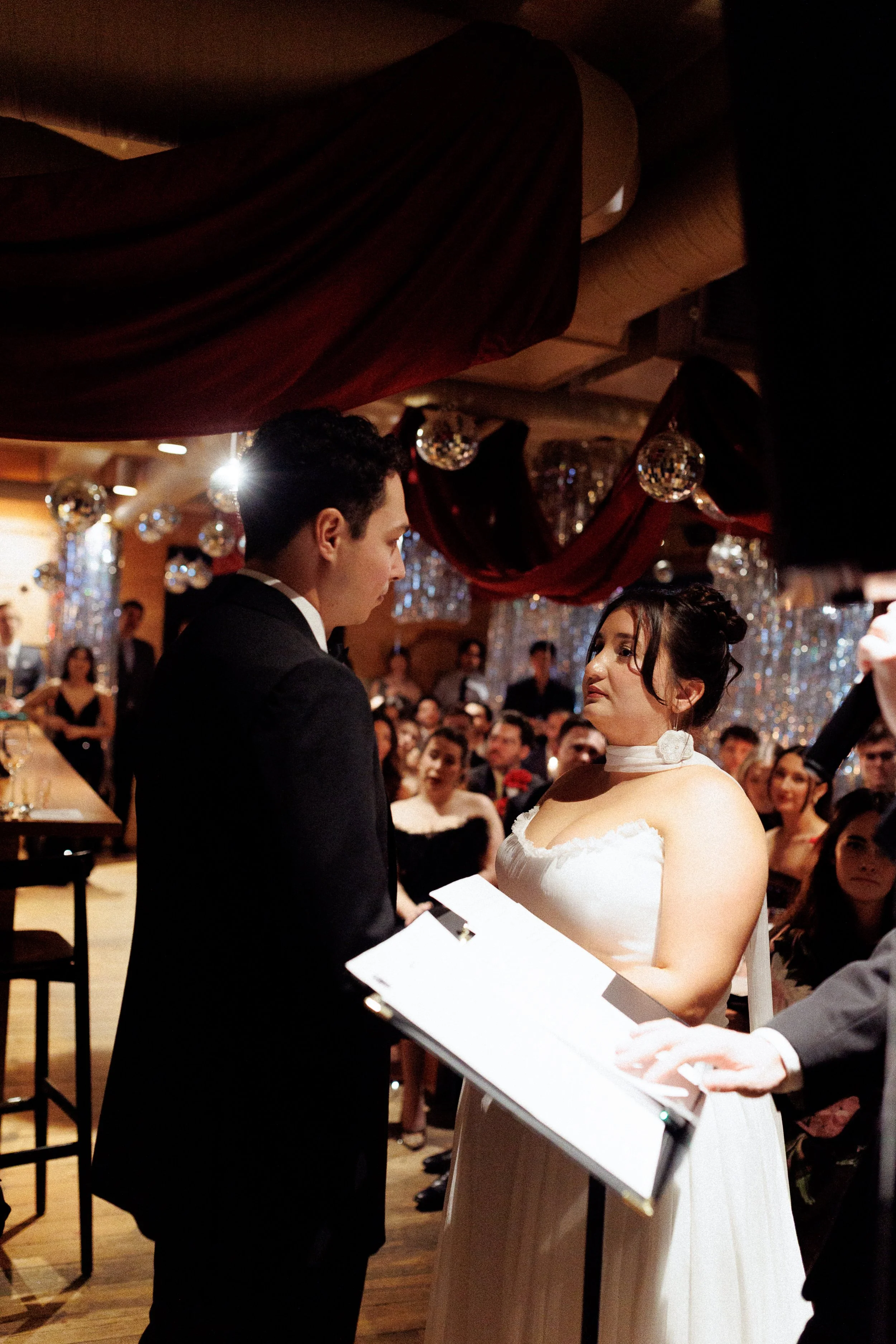 A woman in a white wedding dress and a man in a black tuxedo stand facing each other during a wedding ceremony, with an audience seated behind them under decorated ceilings with red drapes and disco balls.
