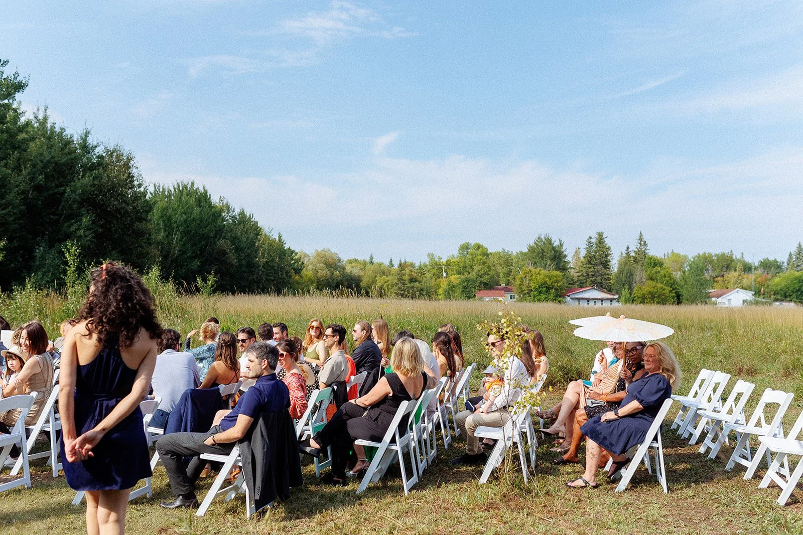 People seated outdoors on white folding chairs during a sunny daytime event in a grassy field surrounded by trees.