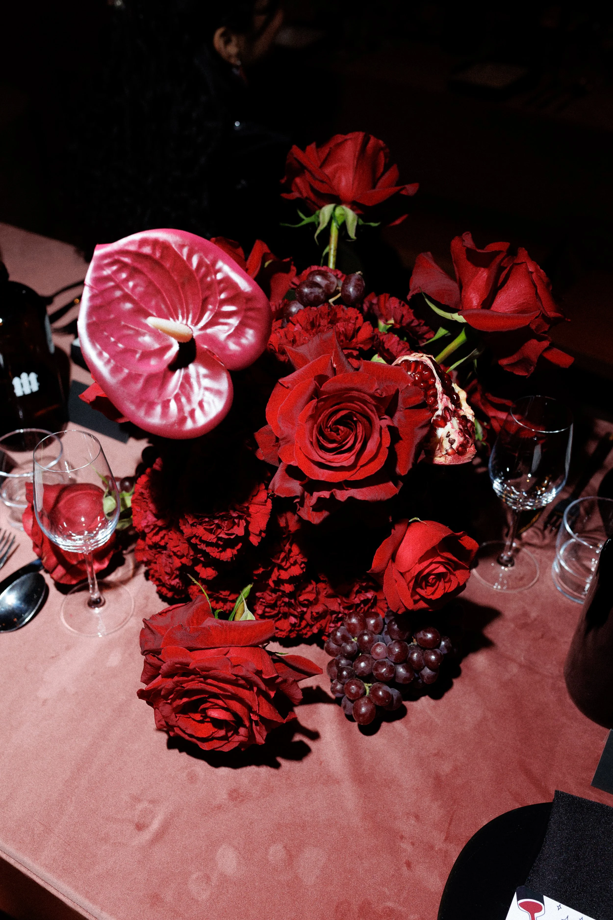 A floral table centerpiece featuring red roses, dark red carnations, grape clusters, and a pink anthurium, set on a pink tablecloth with empty wine glasses around.