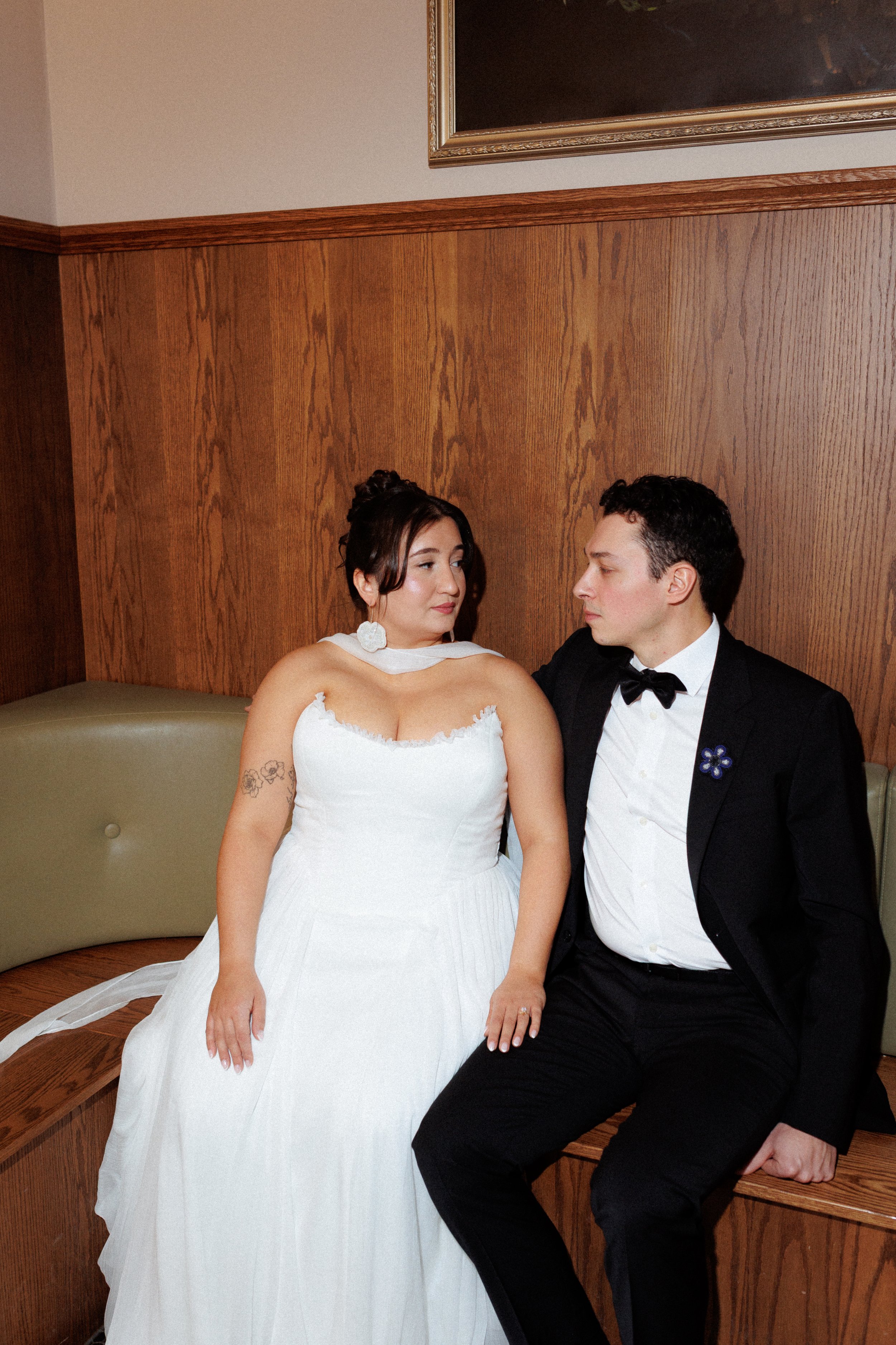 A bride and groom sitting closely on a wooden bench, gazing at each other in a warmly lit room with wood-paneled walls.