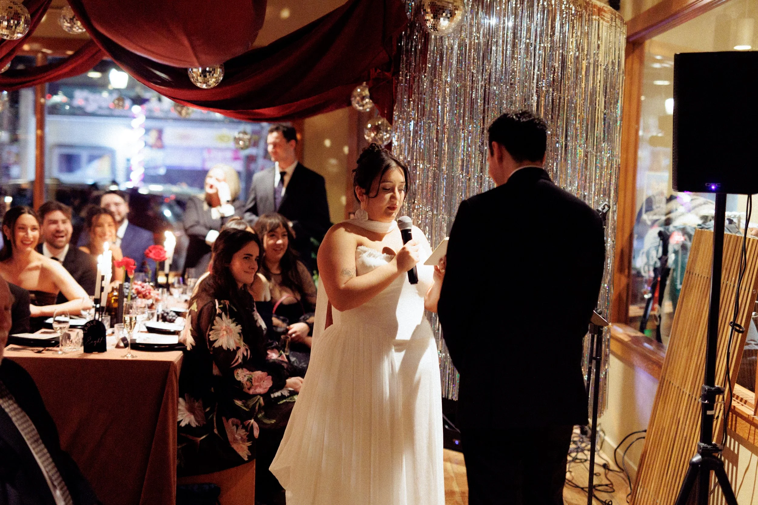 A woman in a white wedding dress reading a vows from a paper, holding a microphone, during a wedding ceremony. A man in a dark suit stands opposite her, with guests seated at tables in the background, watching and smiling.