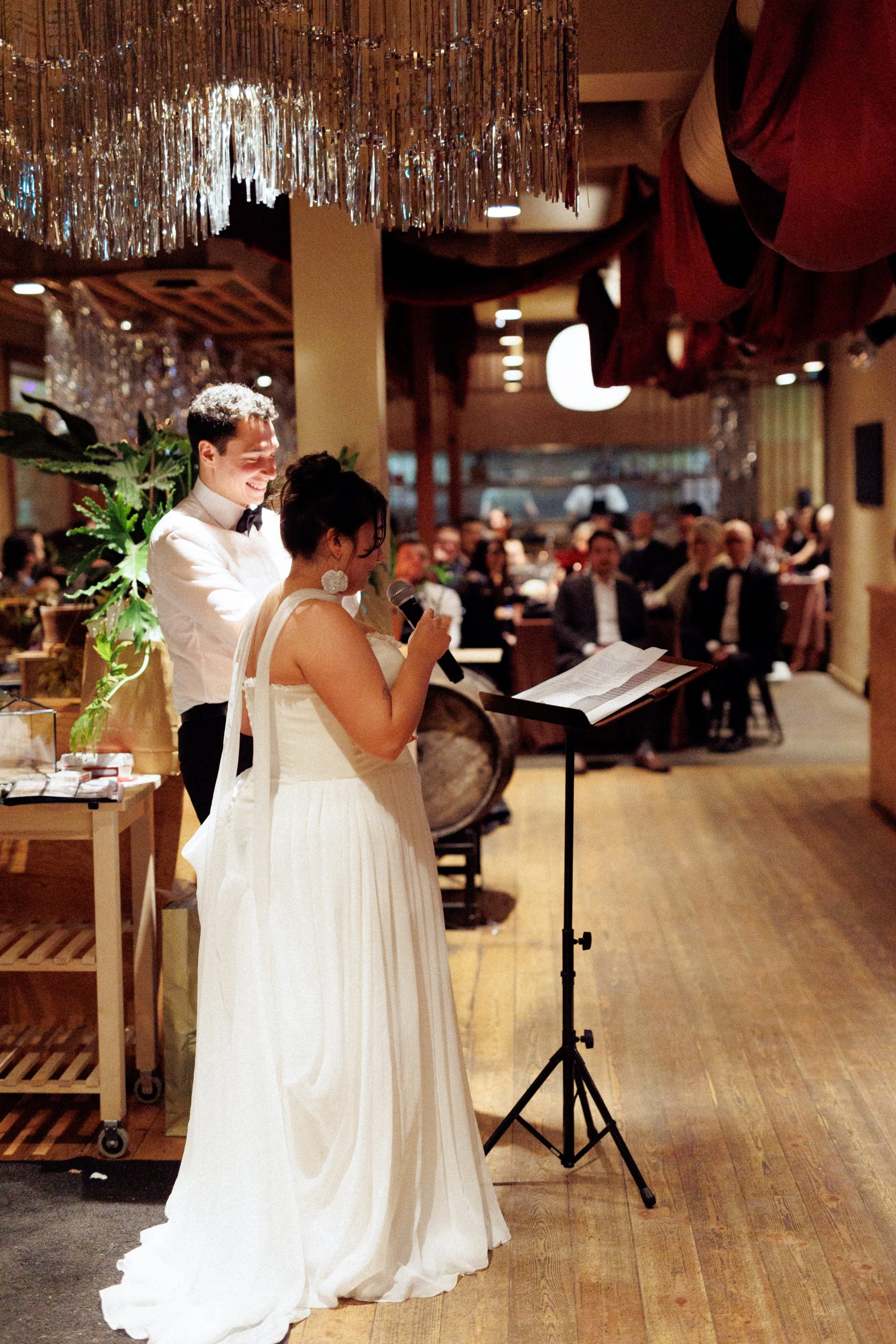 A bride in a white dress and a groom in a white shirt with a black bow tie stand together during a wedding ceremony. The bride is speaking into a microphone, and the groom is smiling behind her. They are in a decorated venue with guests seated at tab
