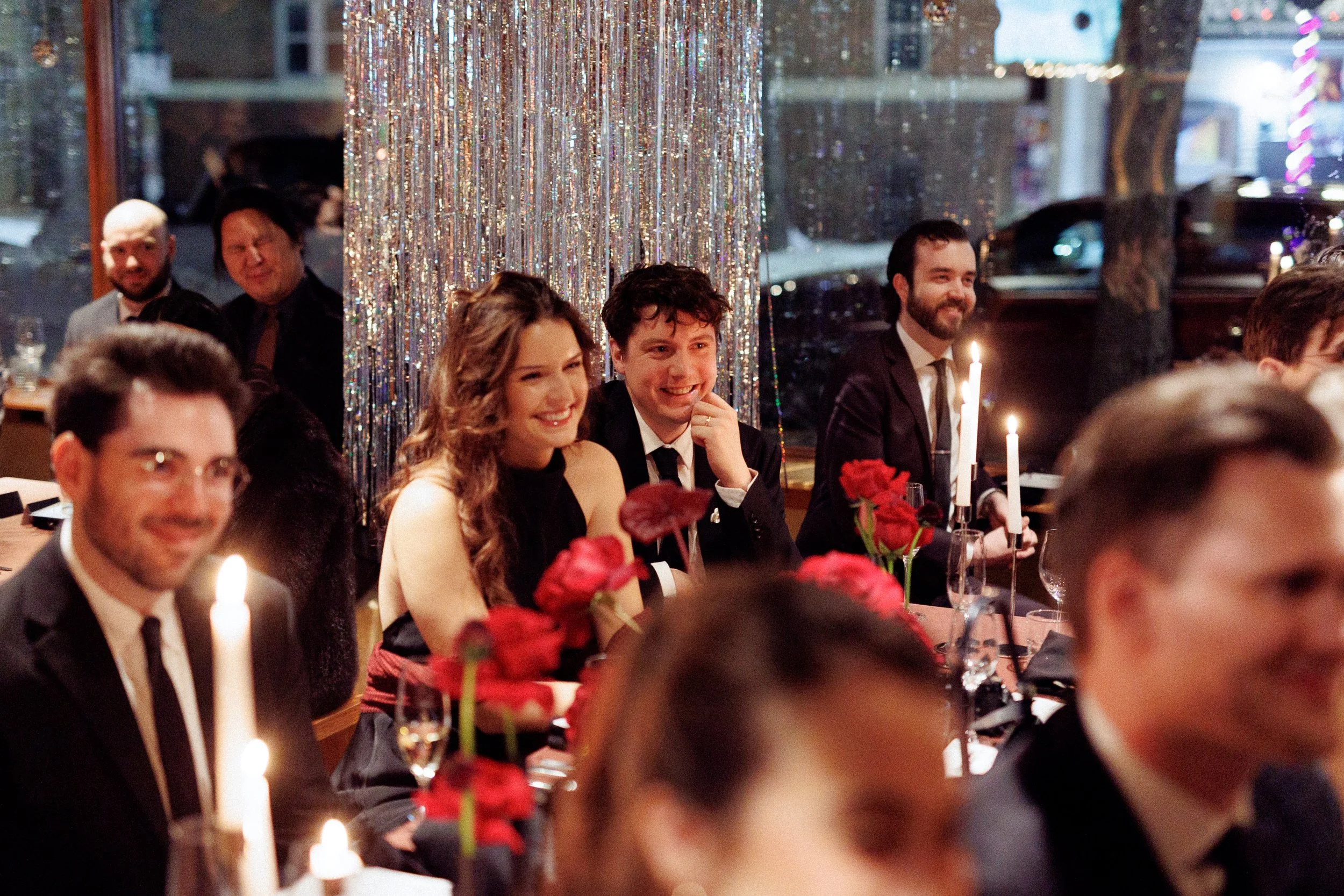 People sitting at a decorated dinner table during a celebration, with some smiling and enjoying the event, candles and flowers on the table, and a shiny, sparkly backdrop.