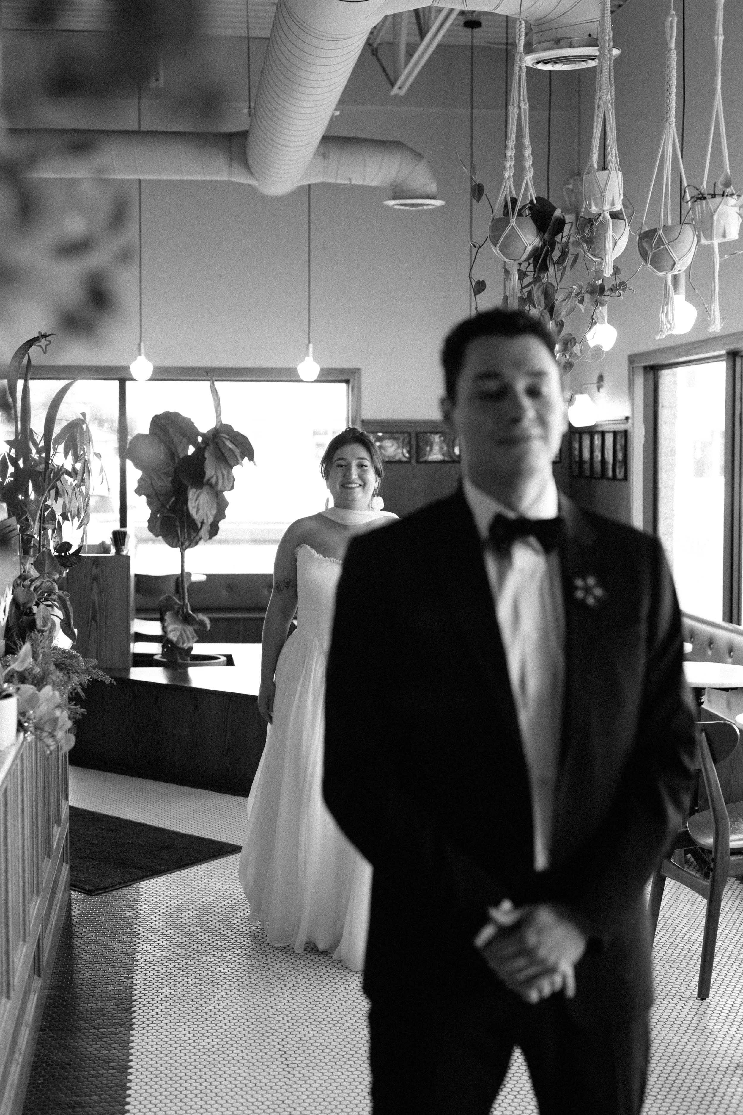 A wedding couple, with the bride smiling in a white wedding dress and the groom in a tuxedo, standing indoors in a decorated space with plants and hanging lights.