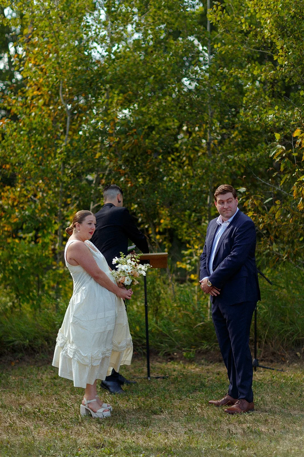 A couple standing outdoors during a wedding ceremony. The woman holds a bouquet of white flowers and wears a light-colored dress with high heels. The man is dressed in a navy suit with a white shirt. In the background, an officiant stands at a wooden