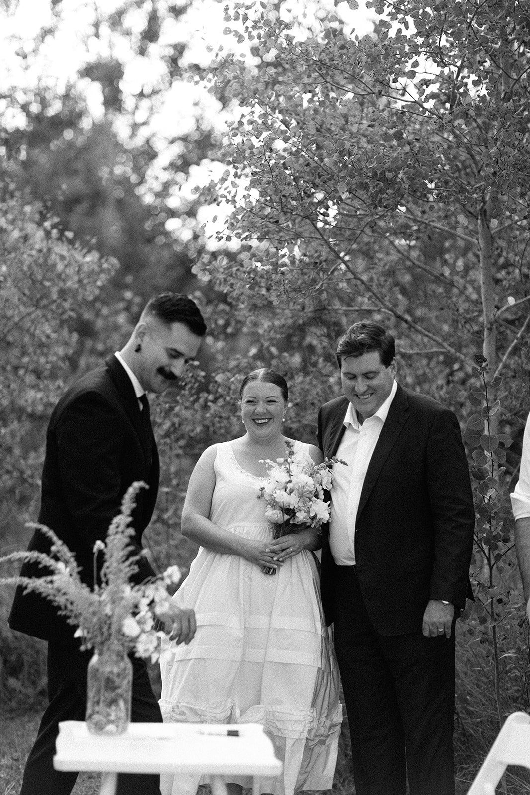 A black-and-white photo of a wedding ceremony outdoors with three people smiling, a woman in a white dress holding a bouquet of flowers, standing between two men in suits, surrounded by trees and foliage.