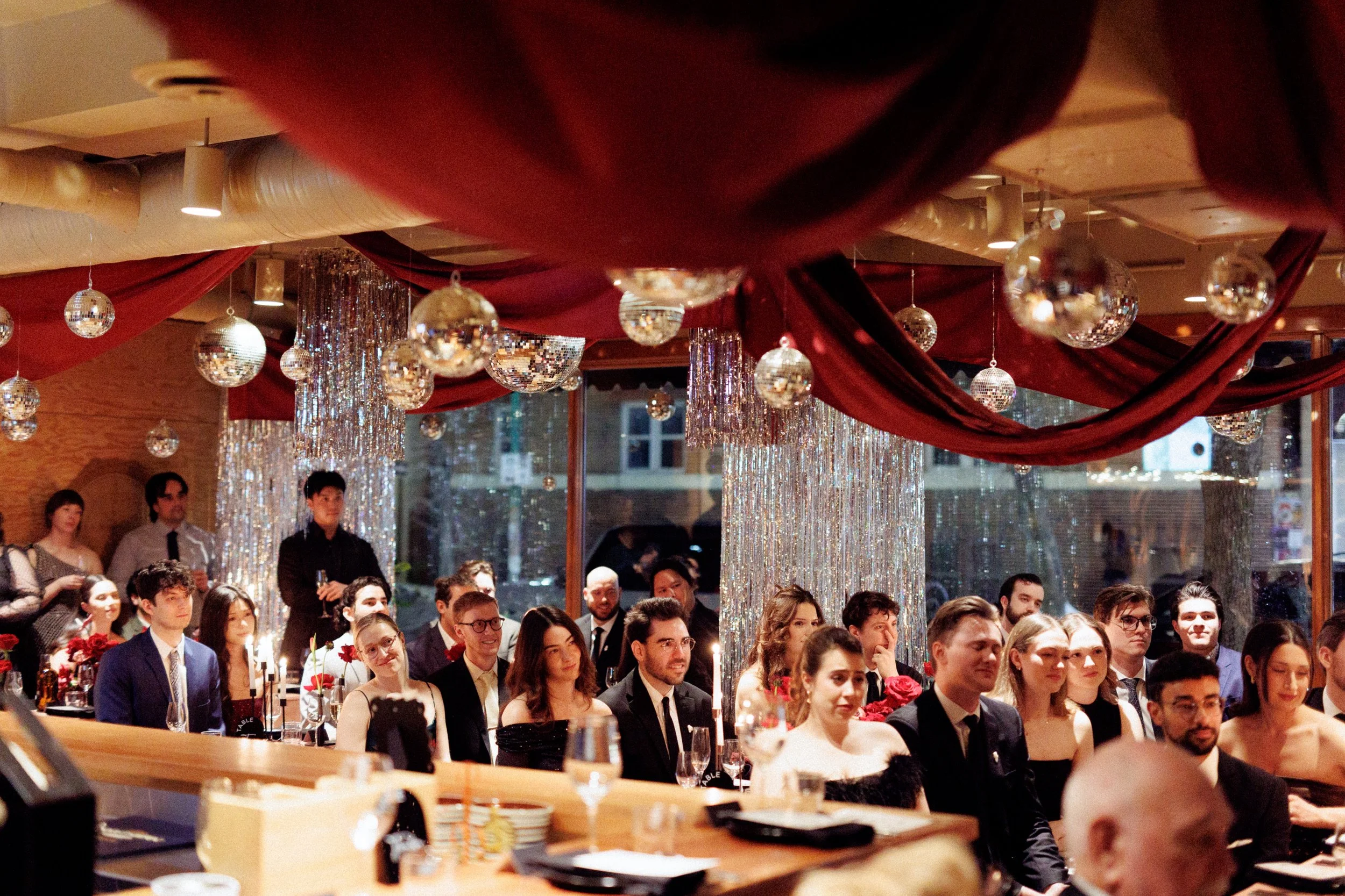 People seated at tables during a formal event or celebration with red draped ceiling and hanging silver and mirror disco balls.