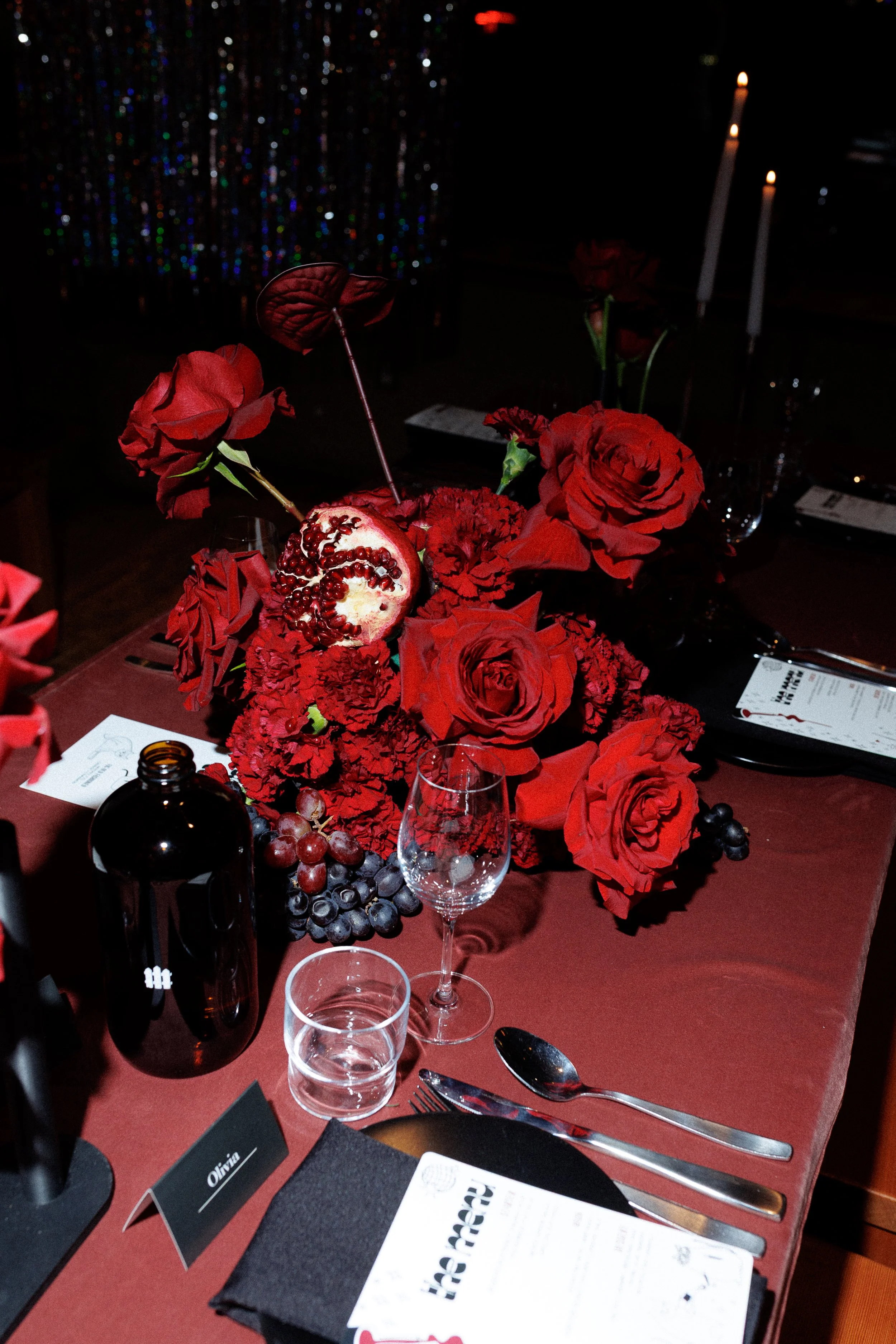 A table setting with a centerpiece of red roses, dark grapes, and a pomegranate half. The table has wine glasses, silverware, a black napkin, and a name card reading 'Olivia'. Candles are lit in the background.
