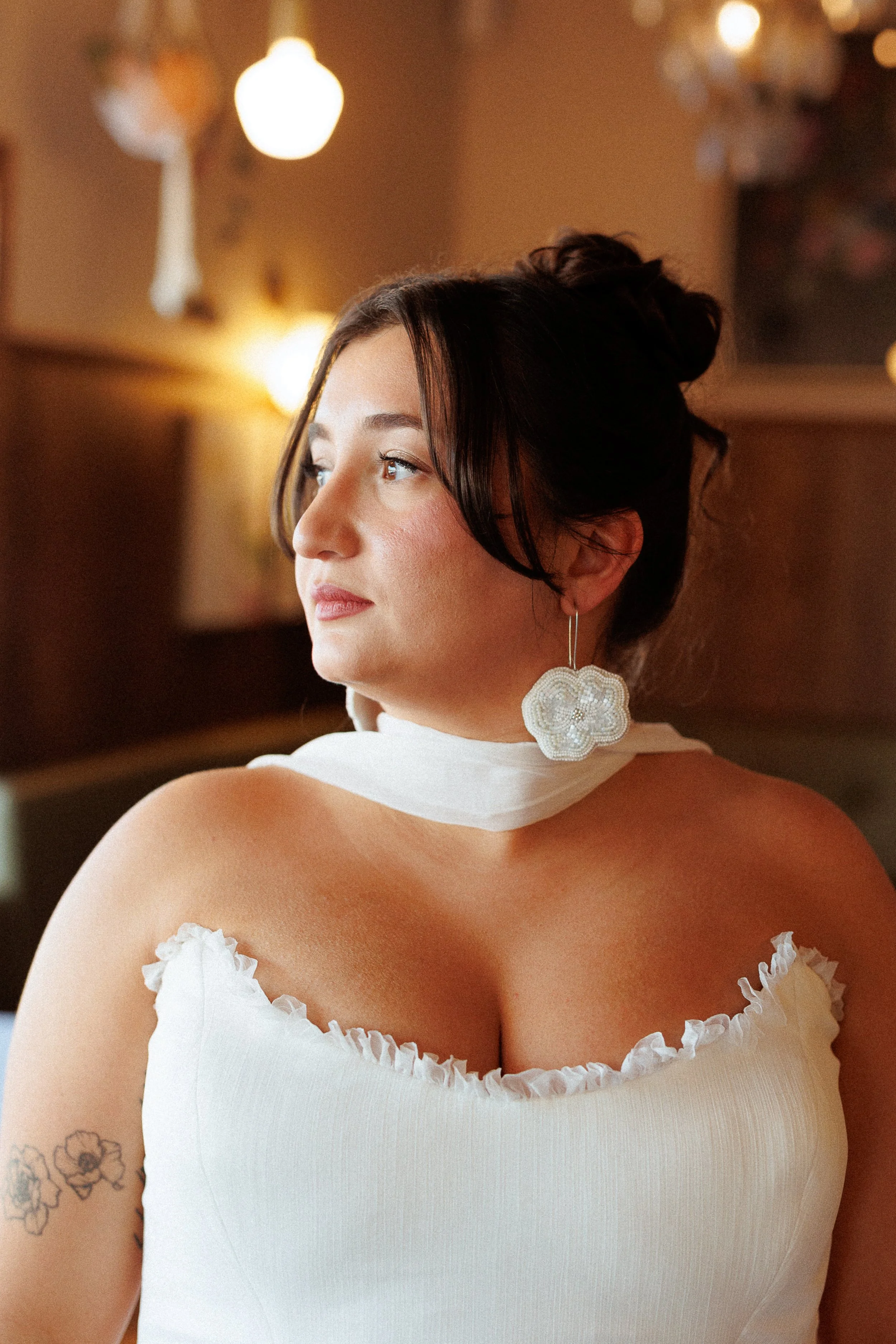 A young woman with dark hair styled in an updo, wearing a white ruffled dress and large flower-shaped earrings, sitting indoors with warm lighting and blurred background.