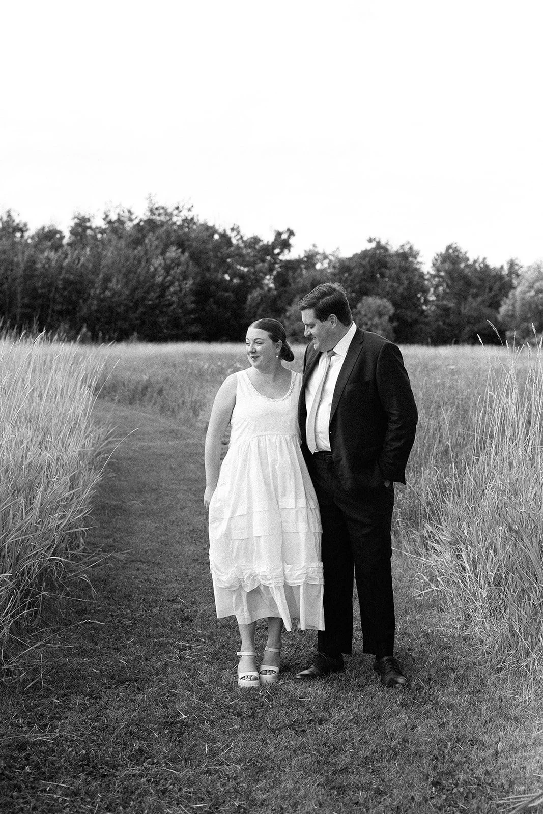 A black and white photo of a woman in a white dress and a man in a suit walking together on a grassy path surrounded by tall grass, with trees in the background.