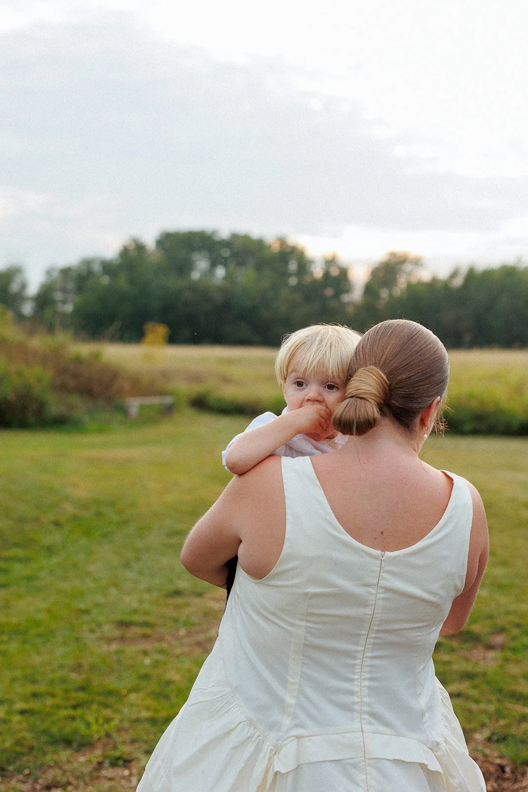 Woman with brown hair in a bun carrying a young blonde boy on her shoulder in a grassy outdoor area with trees in the background.