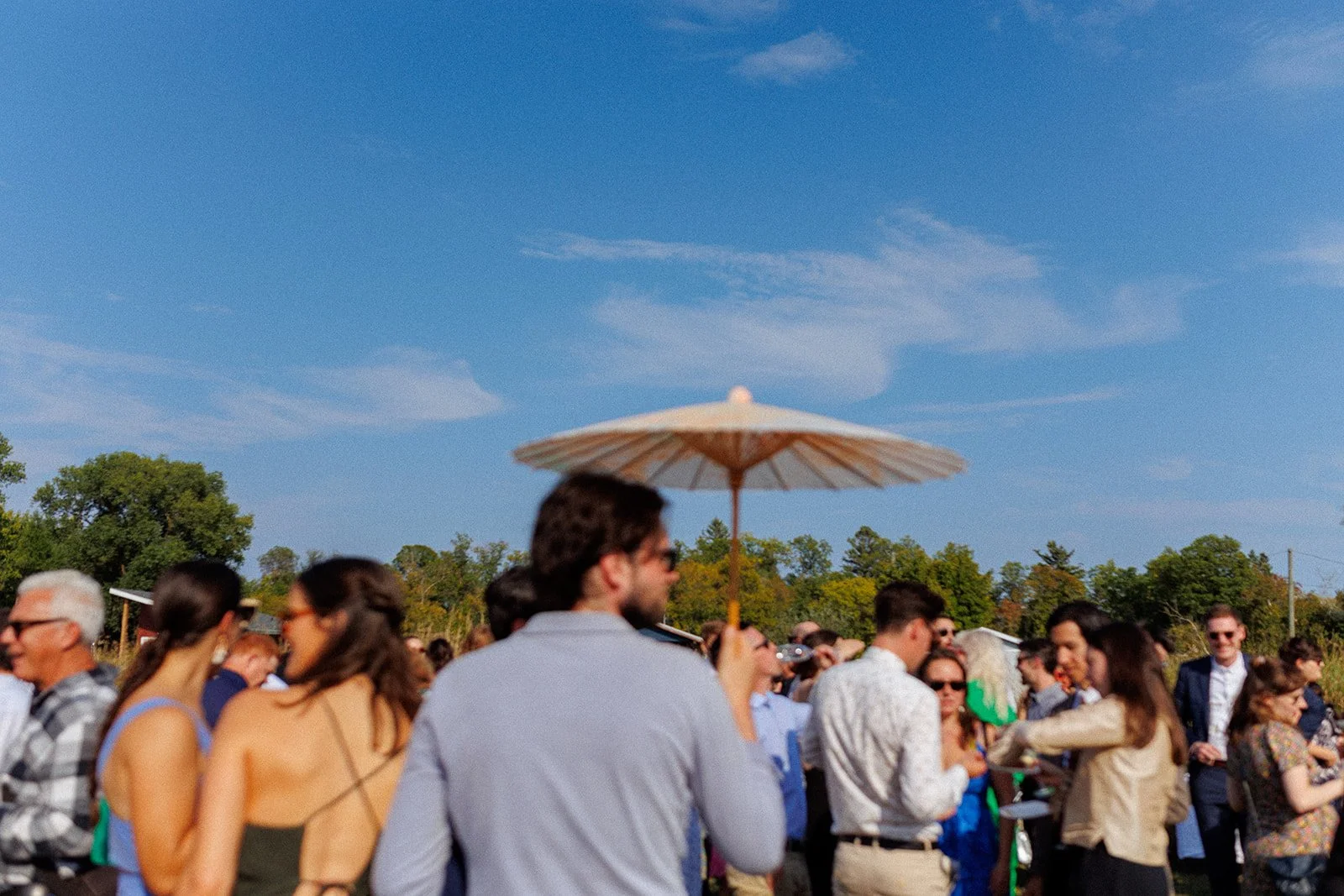 People gathered outdoors on a sunny day, with some holding umbrellas, trees in the background, and a clear blue sky.