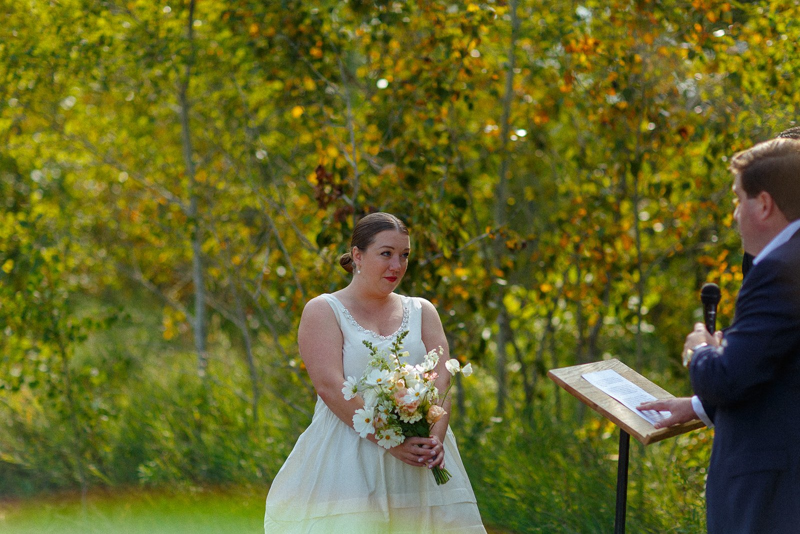 A bride in a white wedding dress holding a bouquet looks at a groom speaking into a microphone during an outdoor wedding ceremony.