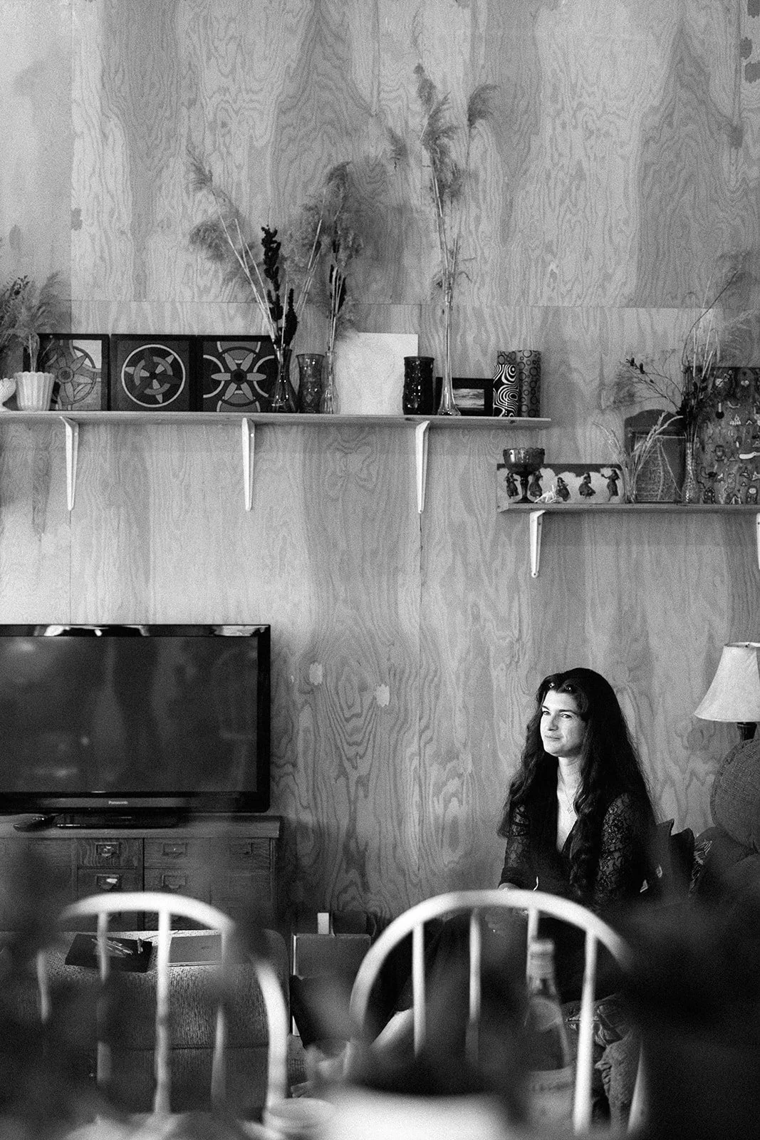 A woman with long dark hair sits at a dining table in front of a wooden wall with shelves holding vases and decorations. A television is visible in the lower left corner.