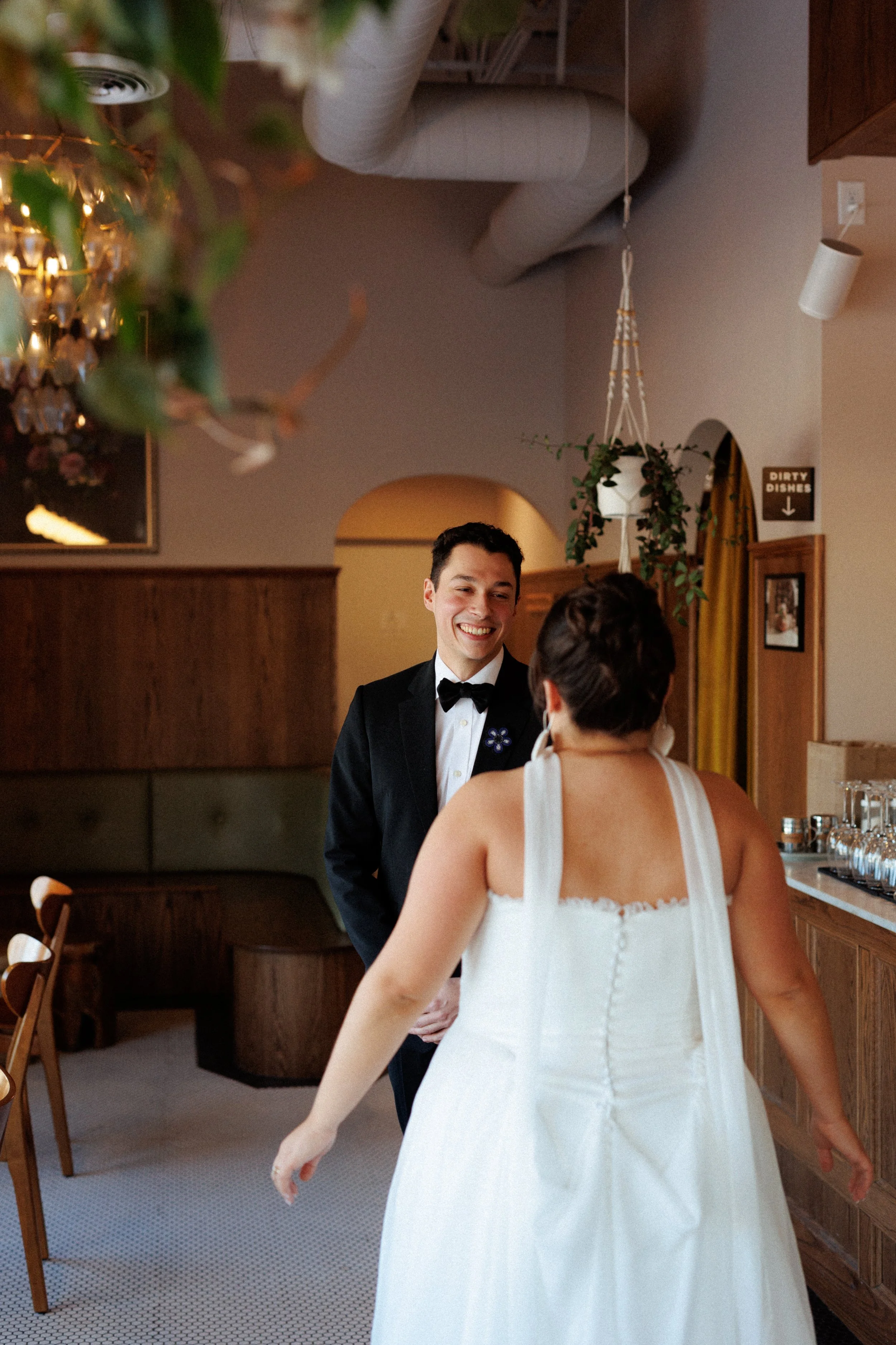A man in a black tuxedo smiling and a woman in a white wedding dress in a cozy restaurant or cafe with warm lighting and wooden furniture.