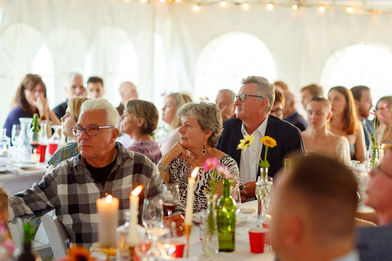 Guests sitting at a long table decorated with flowers and candles during a celebration or event in a bright room with arched windows.