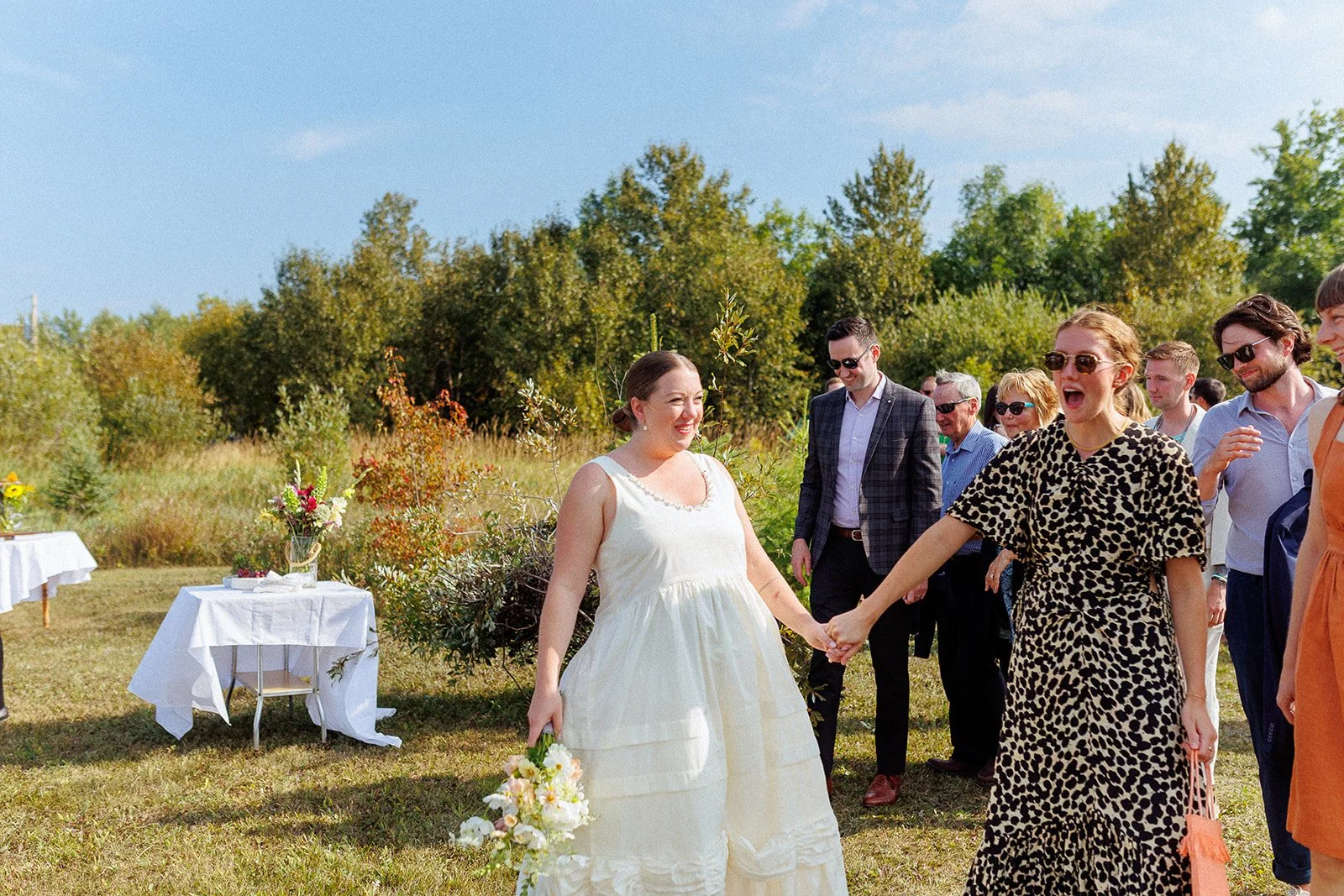 A group of people celebrating outdoors, with a woman in a white dress holding a bouquet and another woman in a leopard print dress holding her hand, smiling and laughing on a sunny day with trees in the background.
