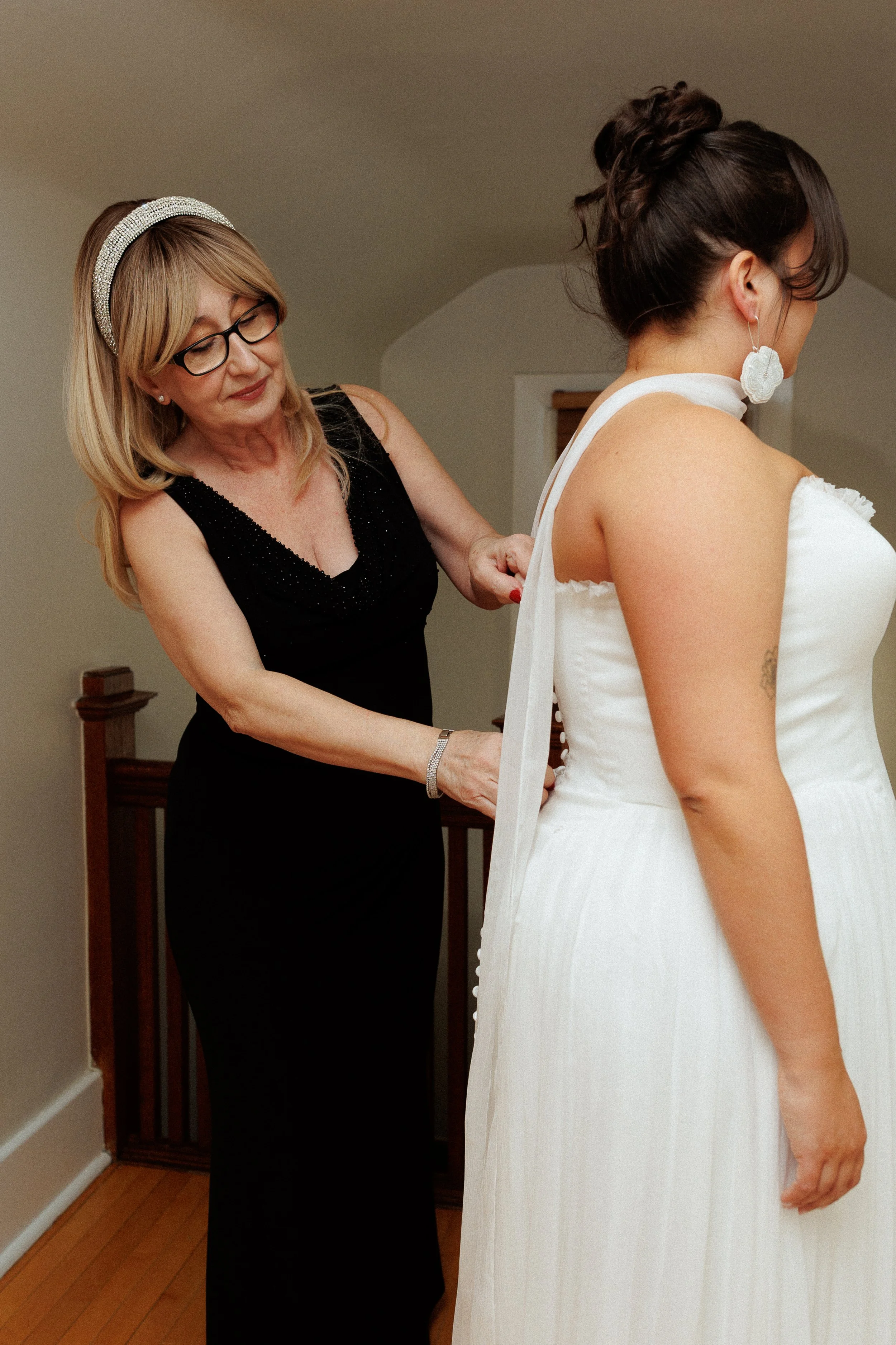 A woman in a black dress helps another woman in a white wedding dress get ready, possibly for a wedding.