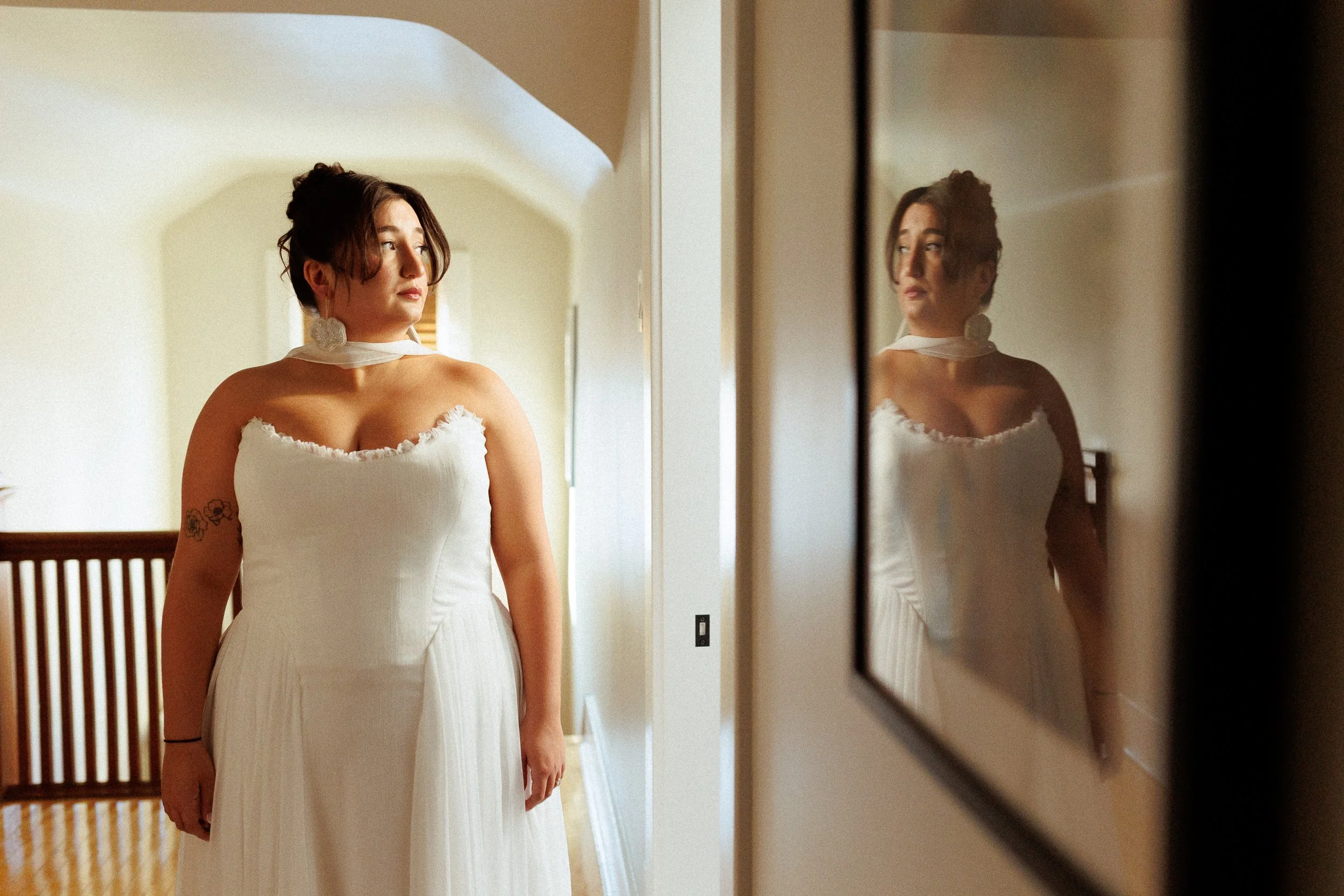 A woman in a white dress standing in front of a mirror at home, with her reflection visible.