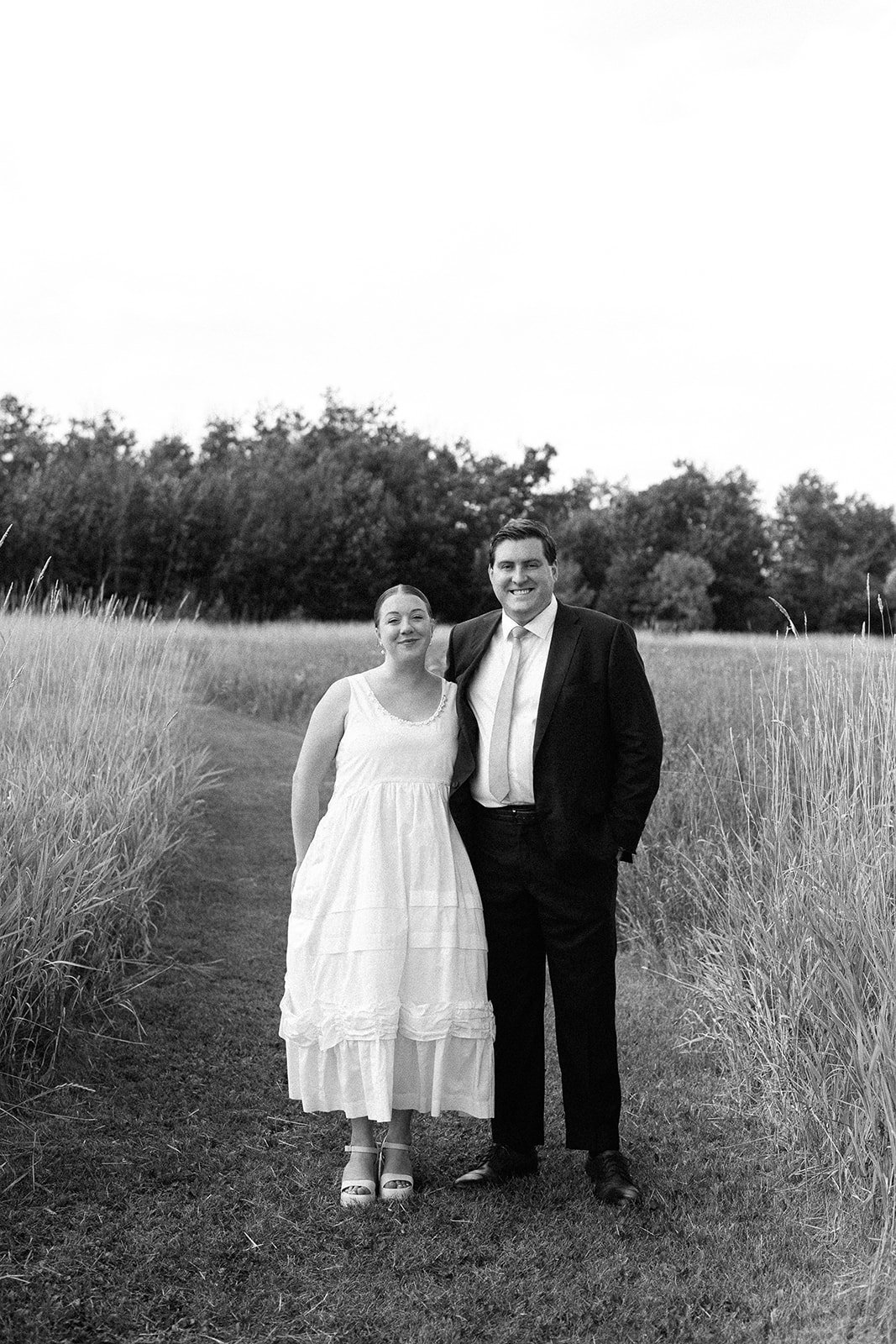 A man and woman stand together outdoors on a grassy path, with tall grass on either side, smiling at the camera, trees in the background, black-and-white photograph.