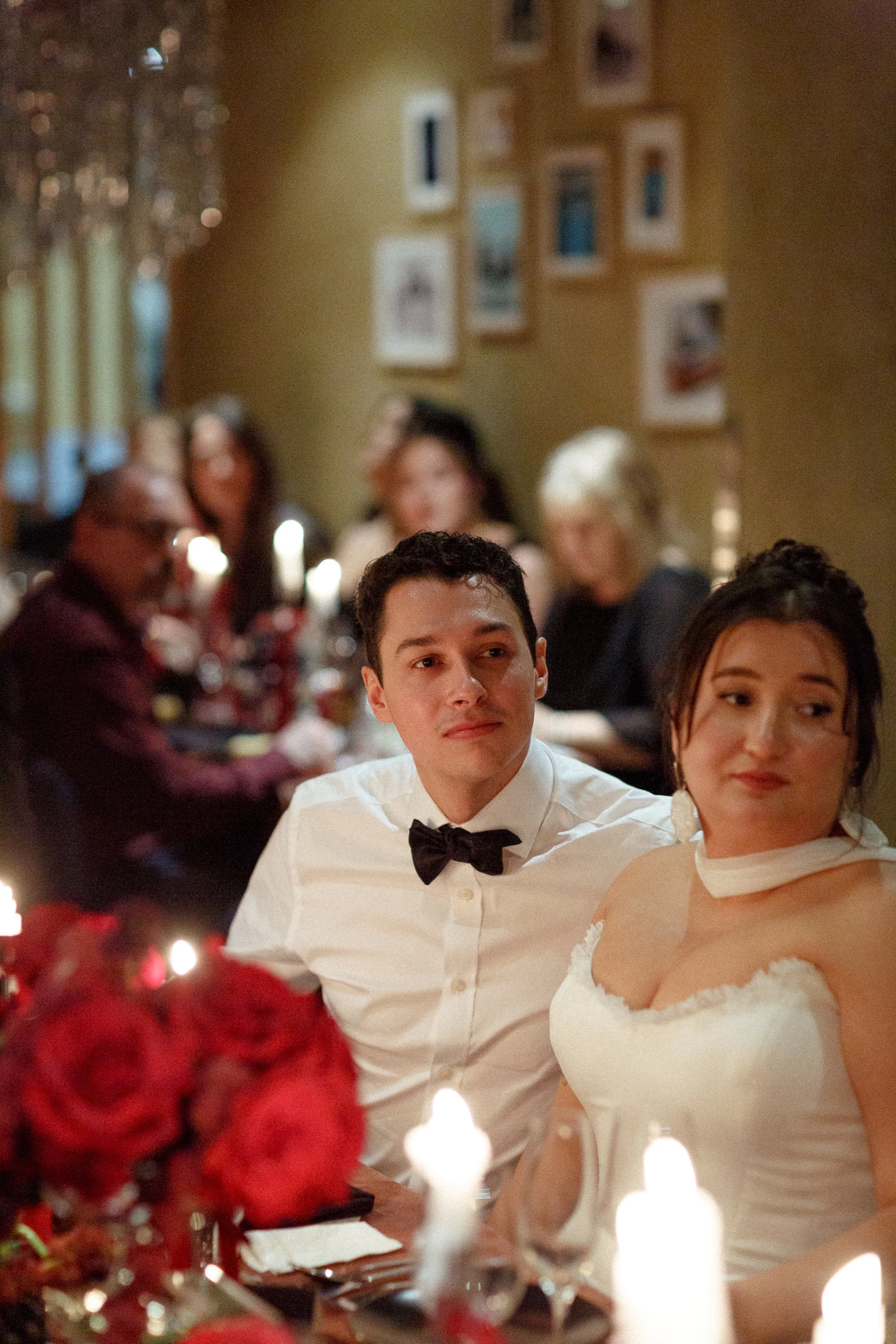 A young man and woman dressed in formal attire sit at a dinner table with candles and red flowers during a wedding celebration, with guests in the background.