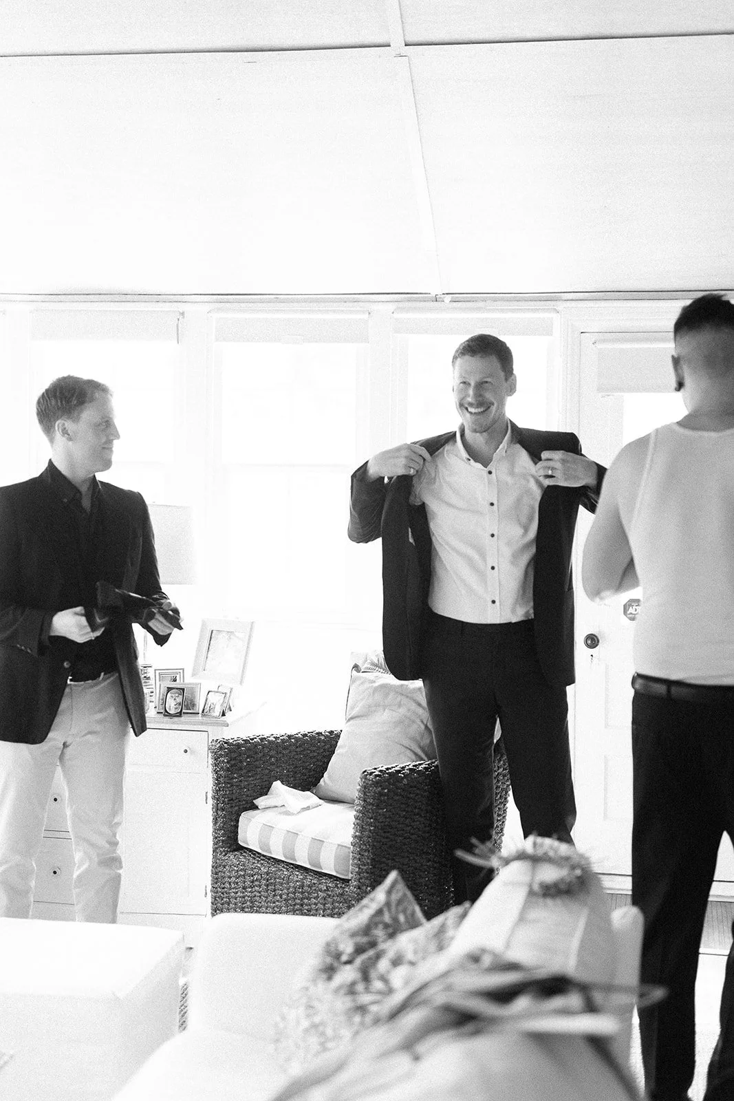 A man in a suit smiling and adjusting his jacket, with two other men in the room, one holding a camera, in a well-lit room with a wicker chair and framed photos in the background.